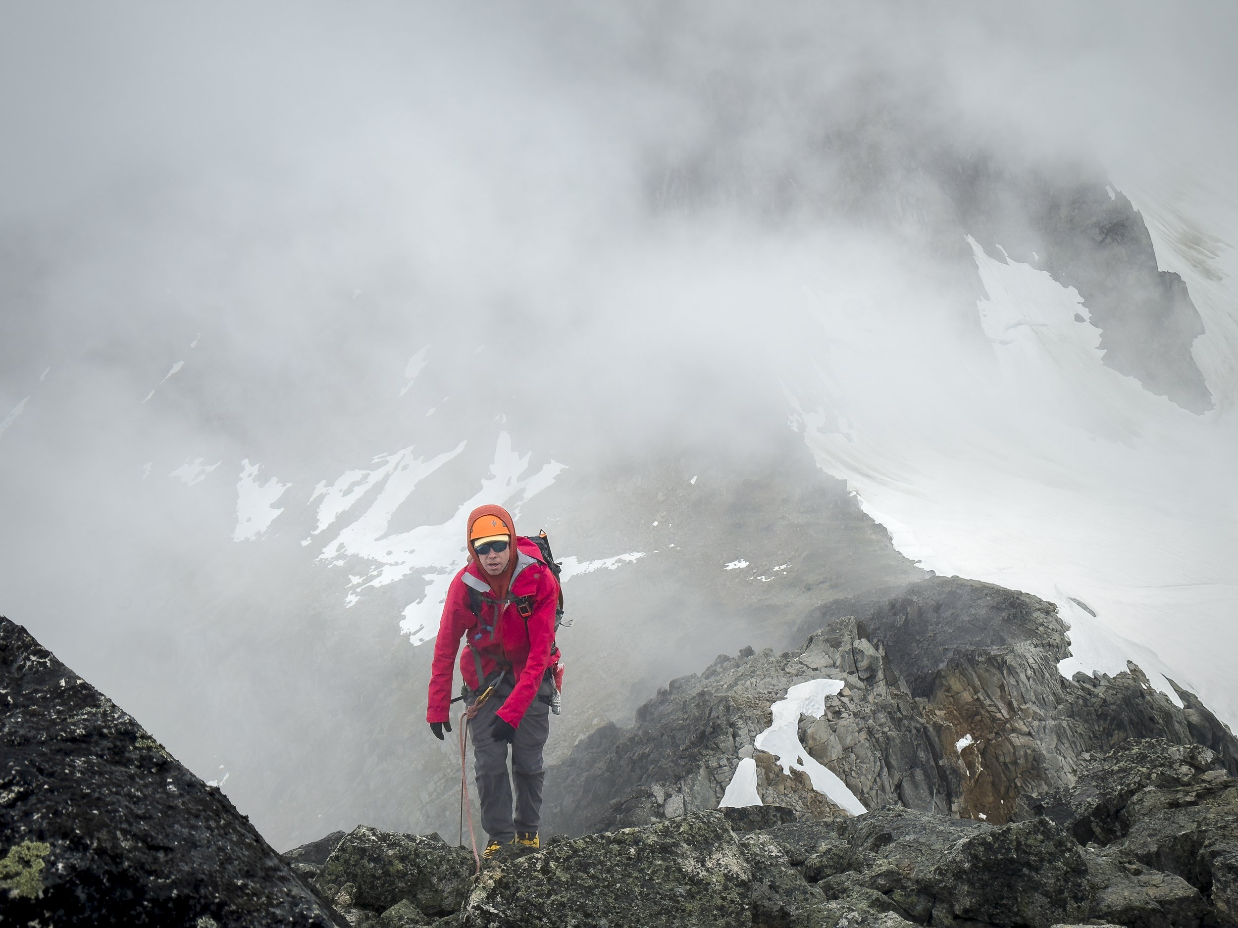 A person in a red jacket, orange helmet, and sunglasses climbing rocky mountain terrain with snow patches and foggy weather.