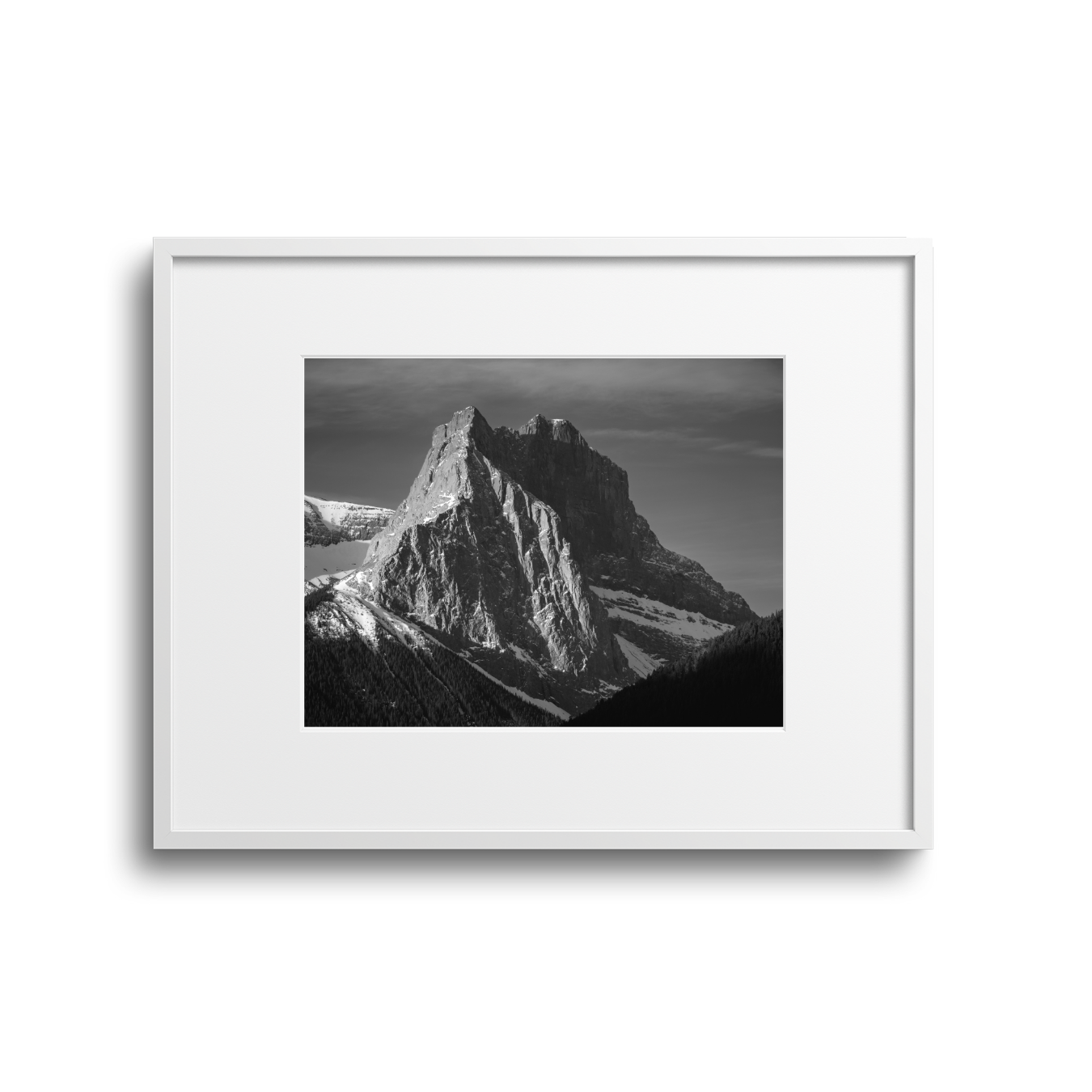 Black and white photograph of a mountain with steep, jagged cliffs and snow patches, framed in a white picture frame against a plain background.