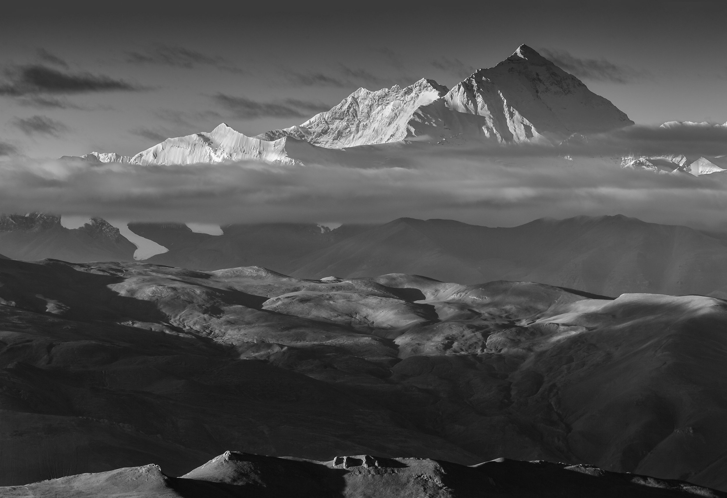 Black and white landscape of snow-covered mountains, with clouds hovering around the peaks and rolling hills in the foreground.