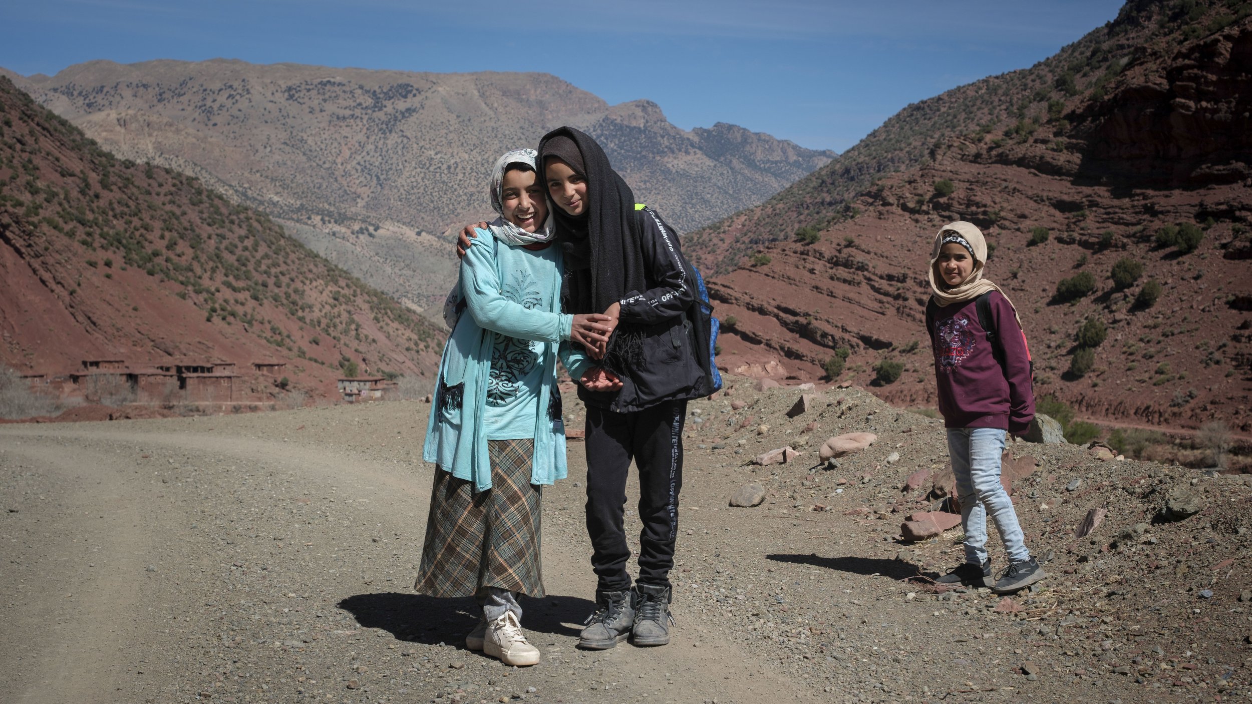 Three children standing beside a dirt road in a mountain valley village in the High Atlas Mountains of Morocco.