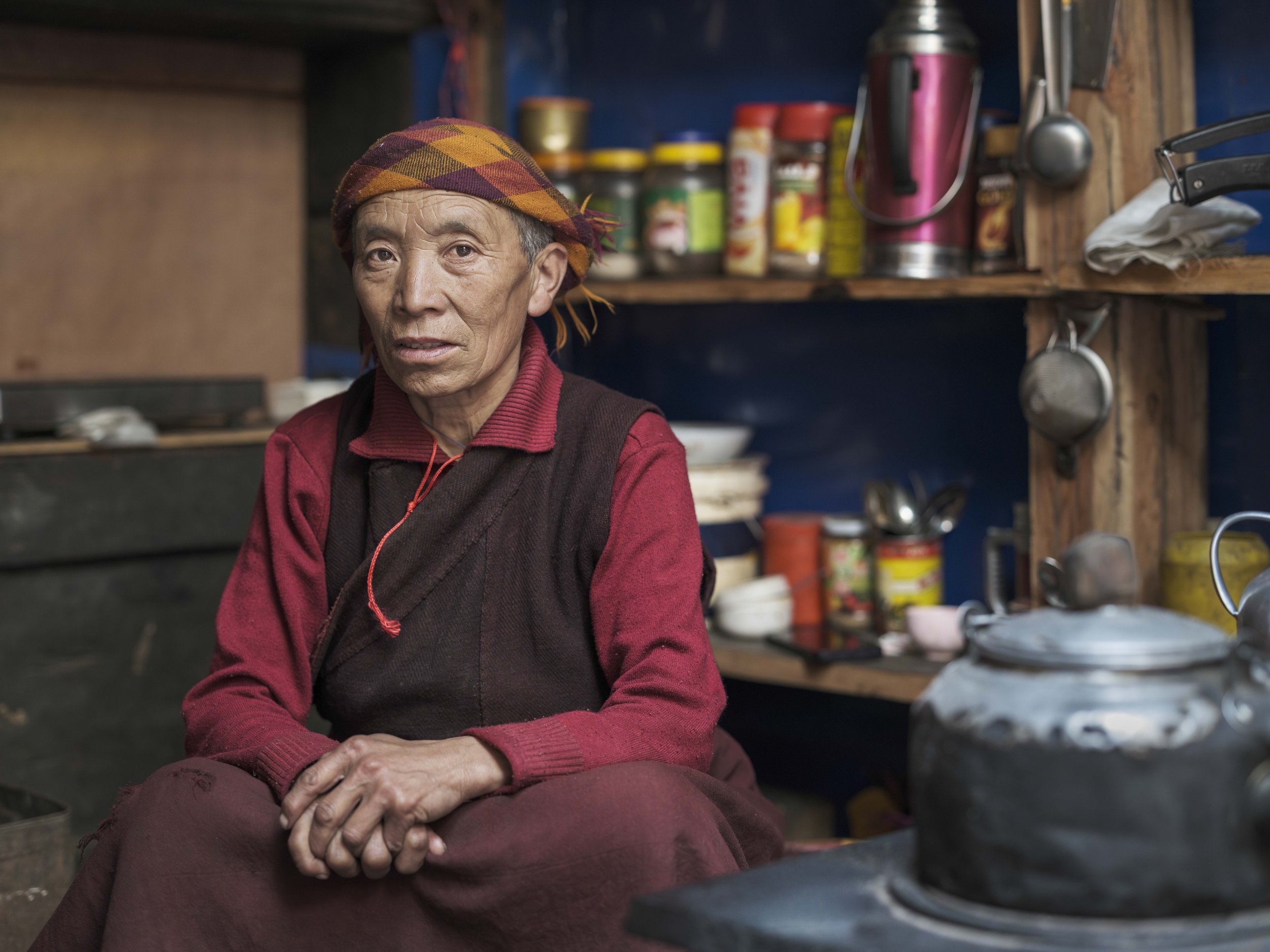 Elderly nun seated inside a traditional kitchen with shelves of utensils and a kettle in Tsum Valley, Nepal.