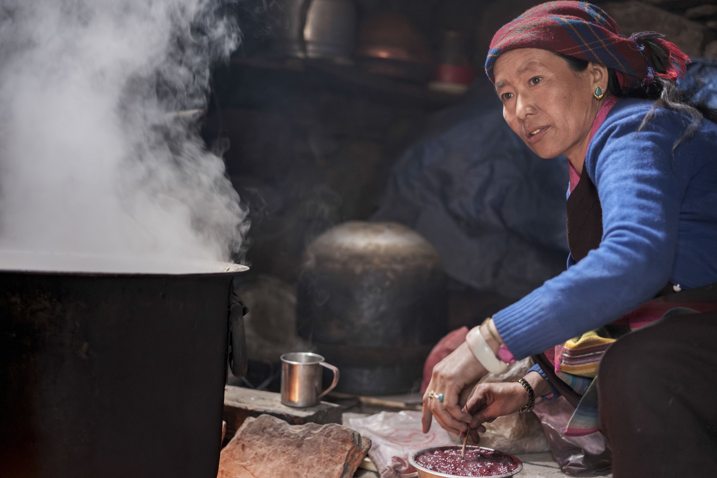 Mingmar tends the pot over the hearth, steam rising in her small kitchen. In Tsum Valley, cloth is often dyed and prepared at home, where fire, wool, and hands shape garments that carry both daily use and tradition. Tsum Valley, Nepal. Photo By Steve