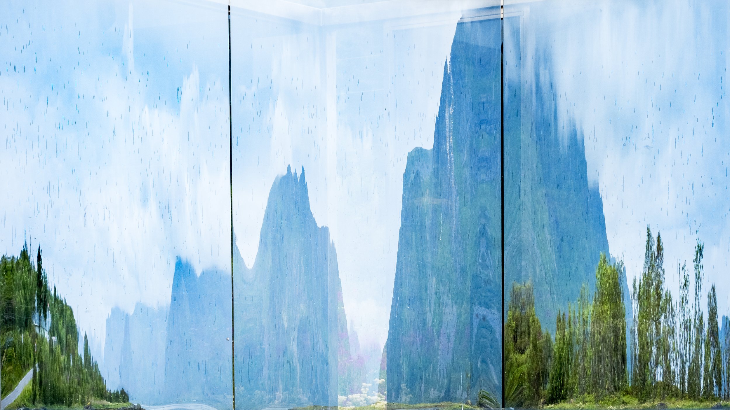 Mountain landscape viewed through reflective glass panels creating layered distortion and fragmented forms.