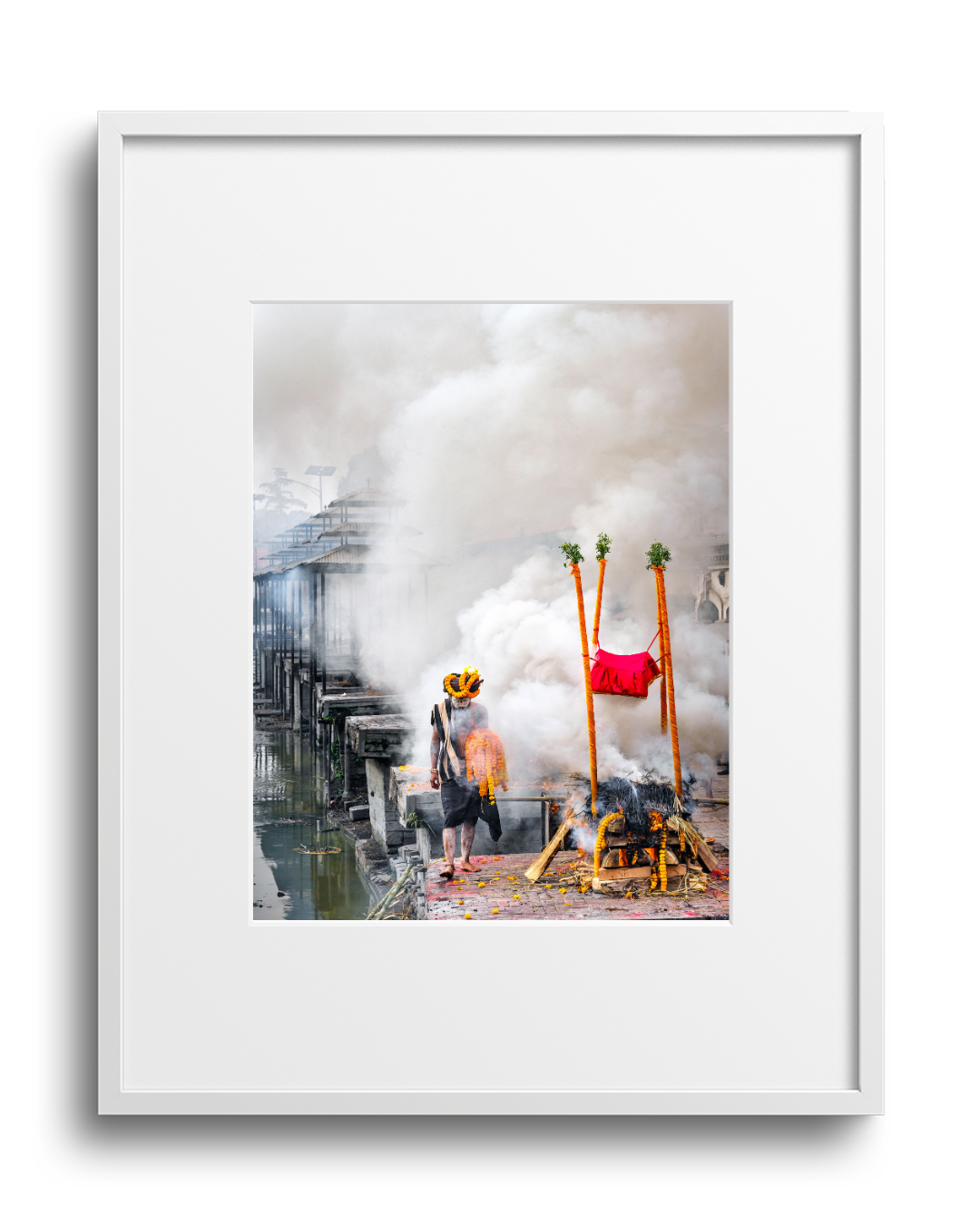 A person participating in a traditional Hindu cremation ceremony on a riverbank, with a funeral pyre ablaze and thick smoke billowing into the sky.