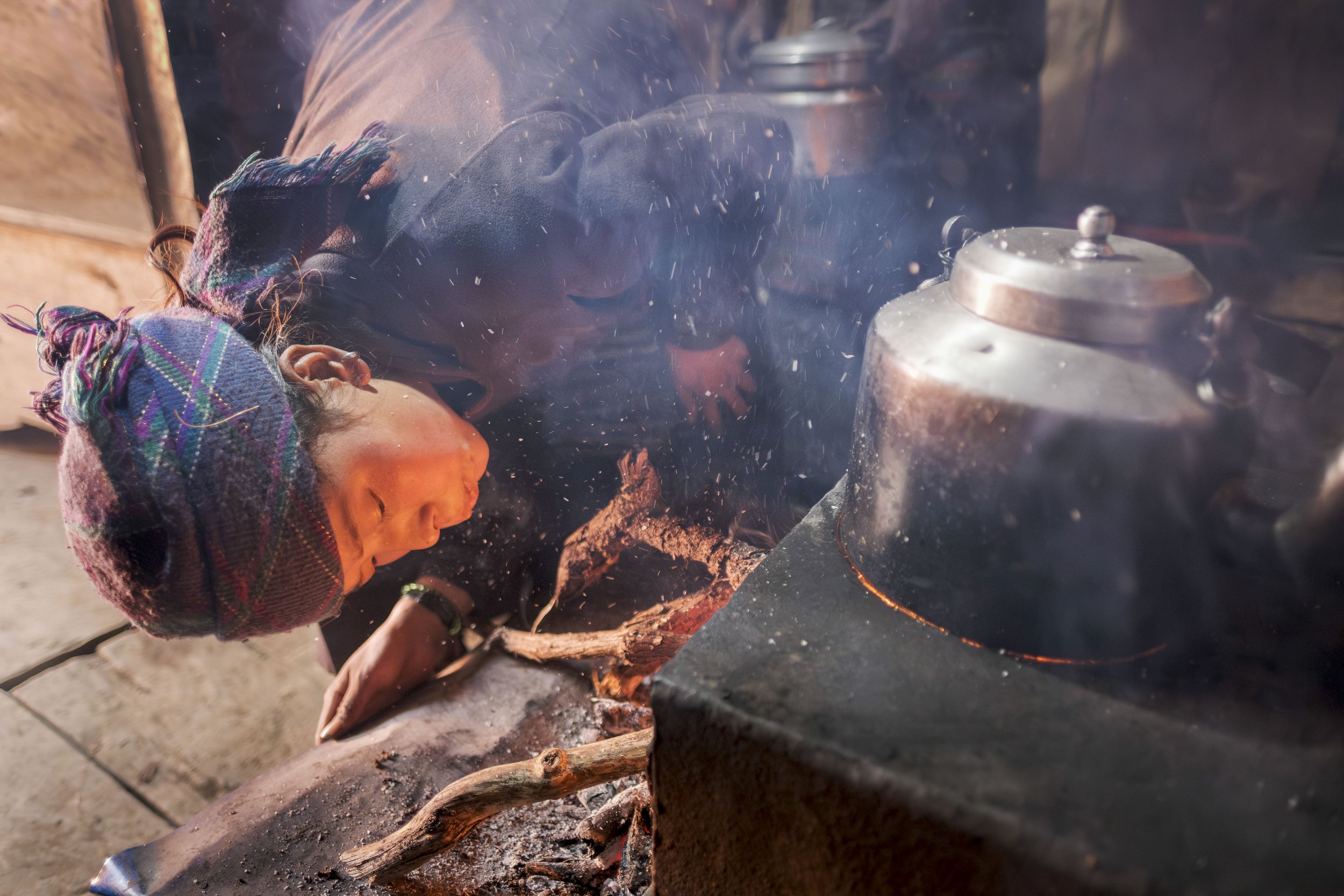 In her kitchen in Chumchet, Sange Lhamu crouches by the open hearth, feeding wood into the fire beneath a blackened kettle, ready to prepare lunch. Tsum Valley, Nepal. Photo By Steve Fagan.