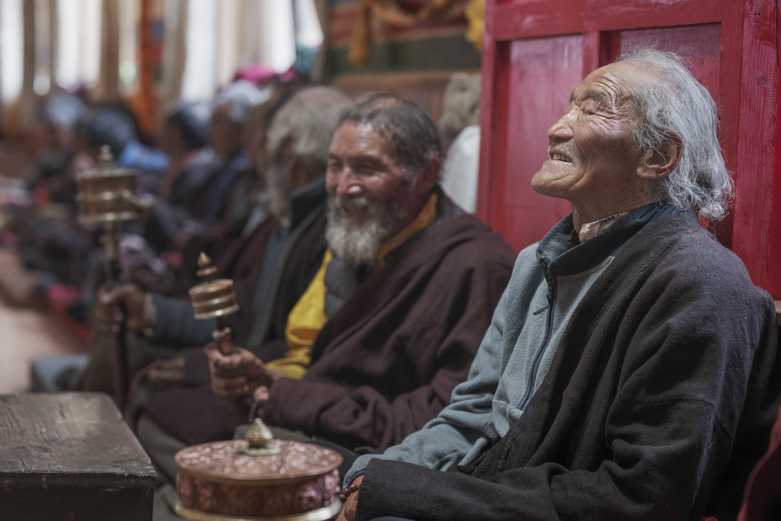 Men sit shoulder to shoulder inside the nunnery hall, sharing laughter mid-prayer, prayer wheels turning steadily. On the first death-anniversary puja of His Holiness Zopa Rinpoche, the valley gathers — farmers, elders, families — holding remembrance