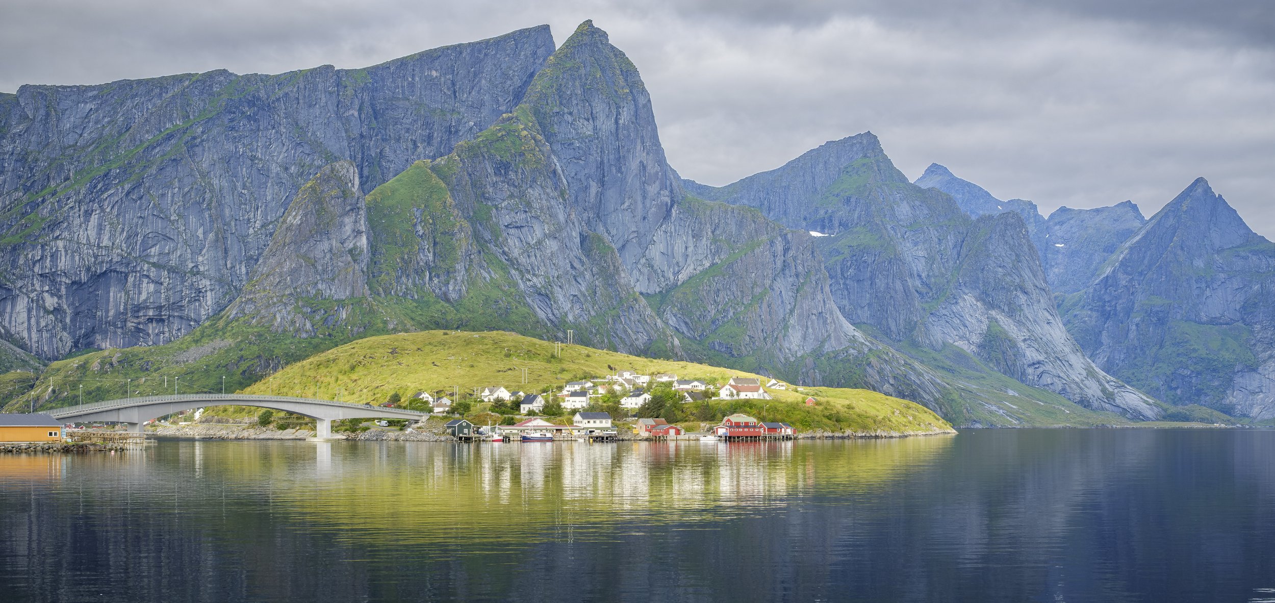 Scenic view of a fjord with water reflecting mountains and a small village with colorful houses. A bridge spans across the water, with a cloudy sky overhead.