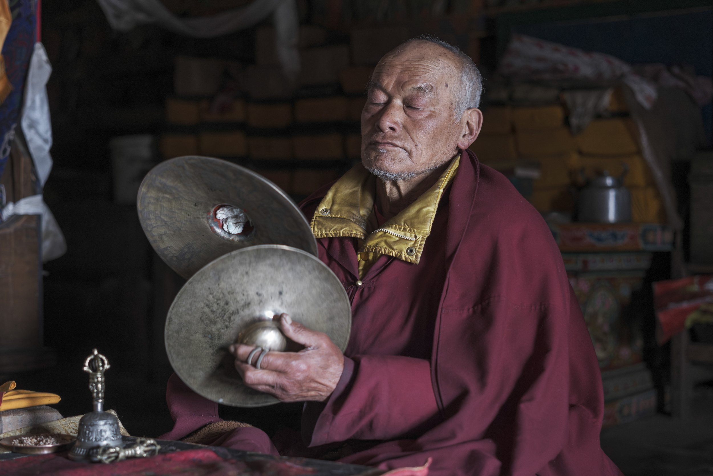 An elderly monk from Mu Gompa sits in concentration, cymbals poised in his hands, eyes gently closed in prayer. With only a few monks remaining, he is among the last caretakers of the monastery's ritual and spirit. Tsum Valley, Nepal. Photo By Steve 