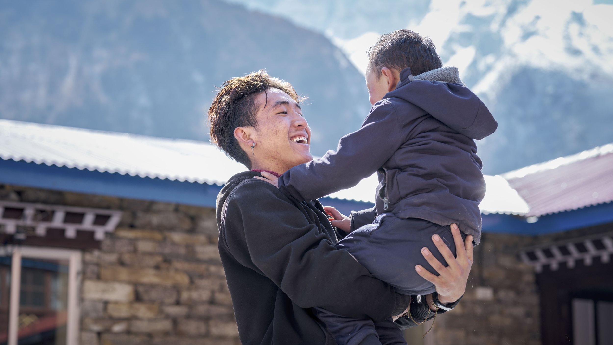 A person with short dark hair and earrings holding a young boy in a dark jacket, outdoors with mountains and building rooftops in the background during daytime.