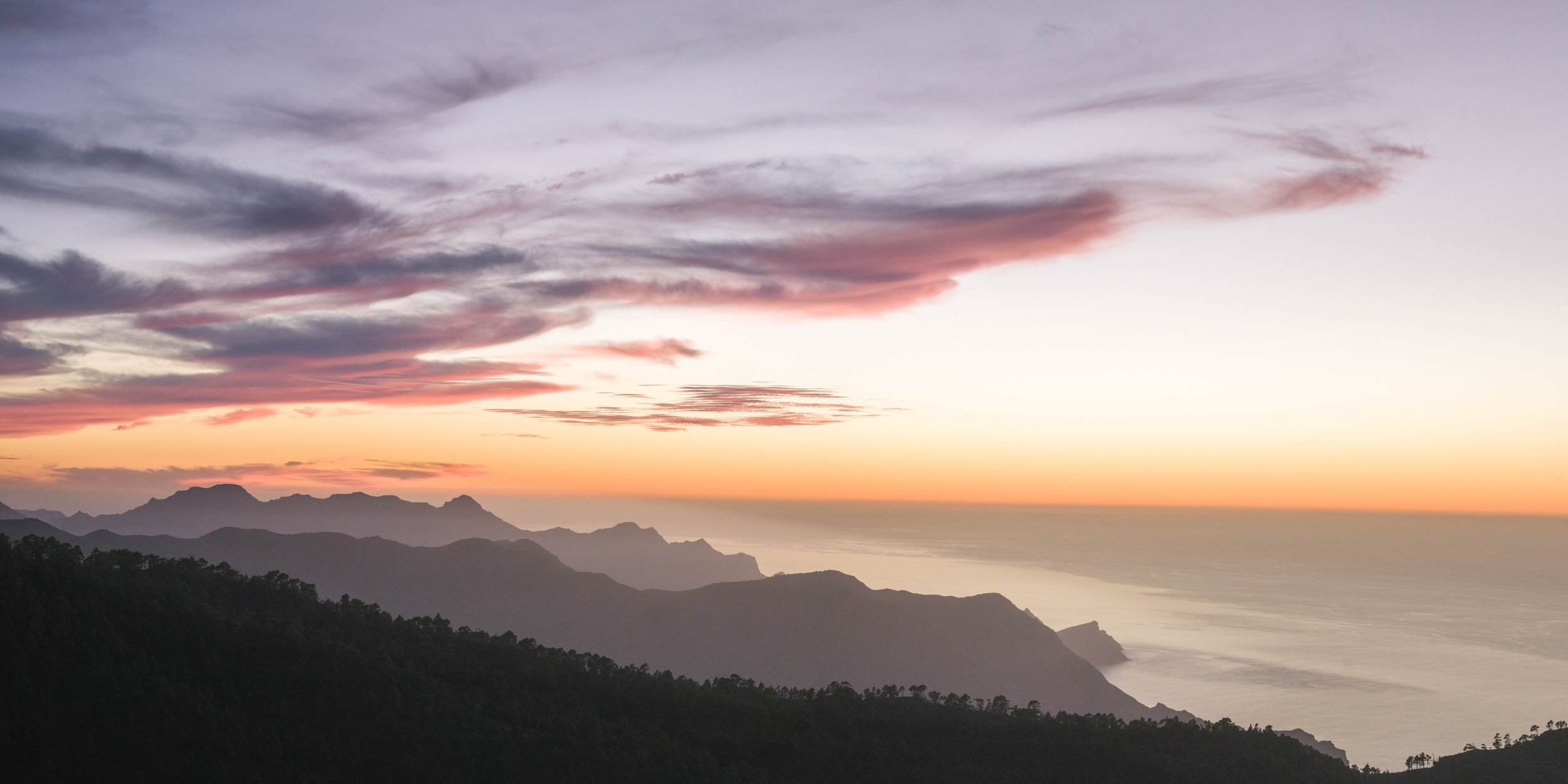 Seaside view of mountains during sunset, with colorful sky and clouds, and the ocean below.