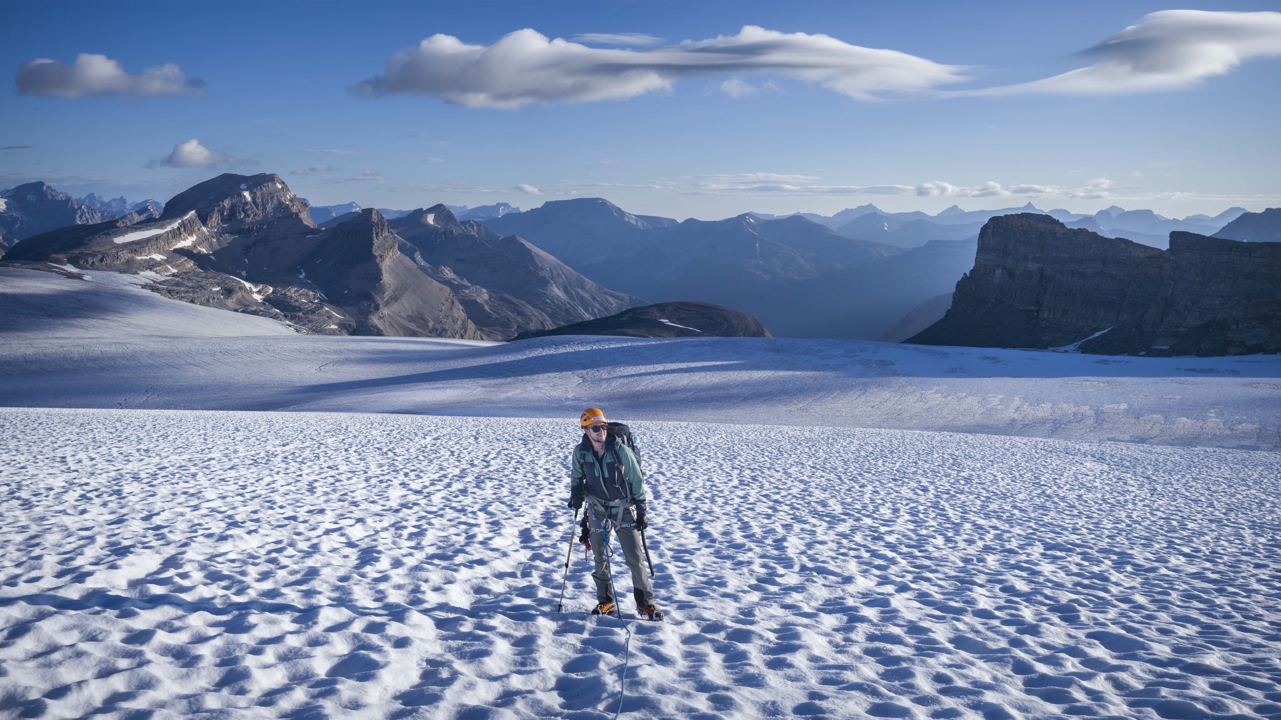 A person in winter gear standing on snow with mountains and a blue sky in the background.