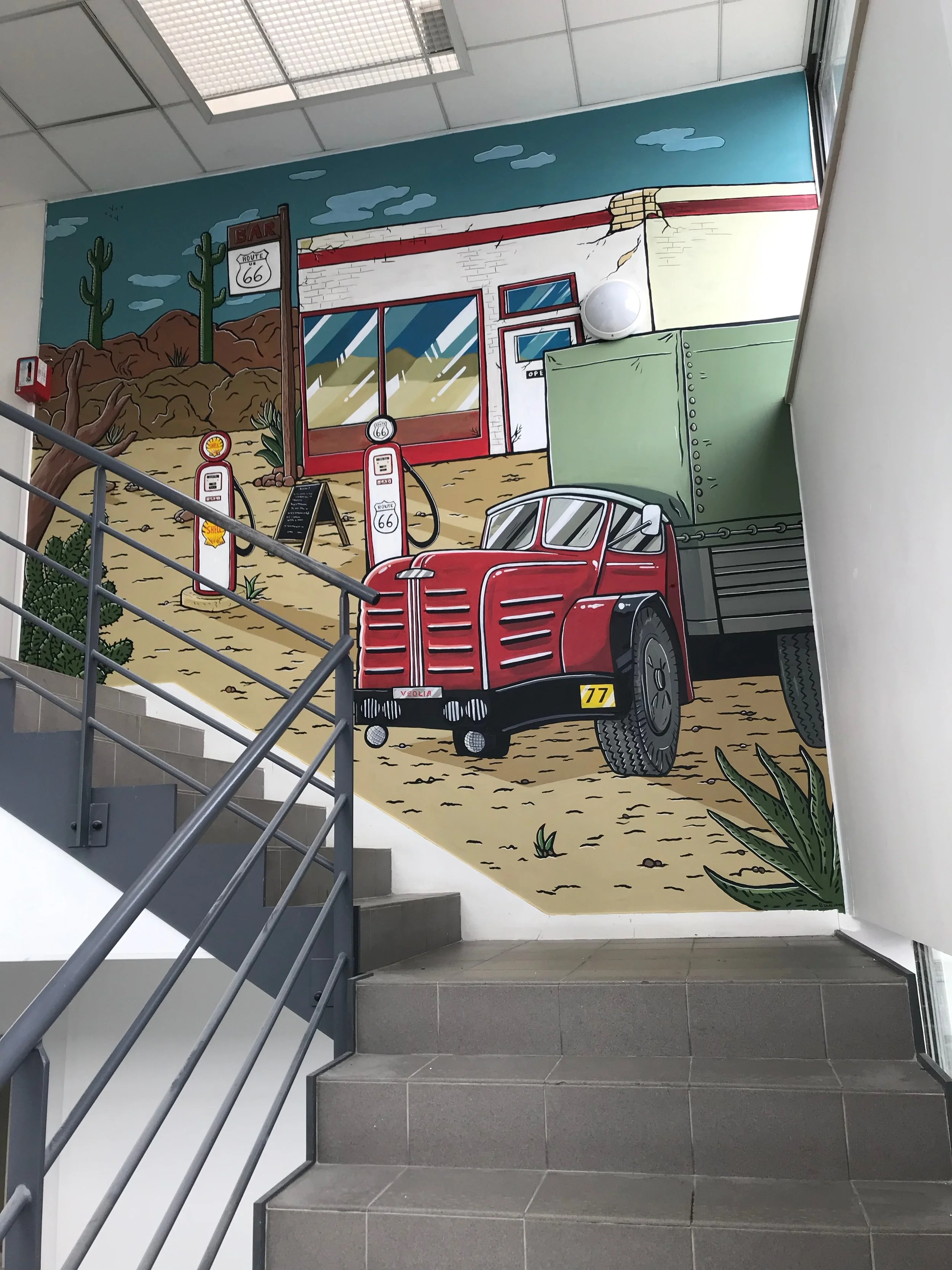 Peinture murale représentant un paysage désertique avec deux cactus, une station essence, un bâtiment avec des fenêtres et une porte, un panneau Route 66, une vieille camionnette rouge, des plantes grasses et un ciel bleu avec des nuages, située à côté d’un escalier en intérieur.
