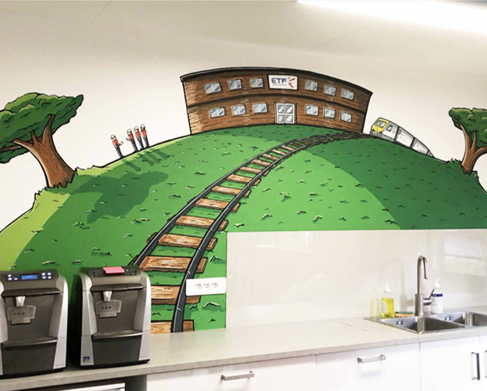 Mural coloré représentant une scène de train et une colline avec deux arbres et un bâtiment, situé au-dessus d'une cuisine avec deux machines à café, un évier et des produits de nettoyage.