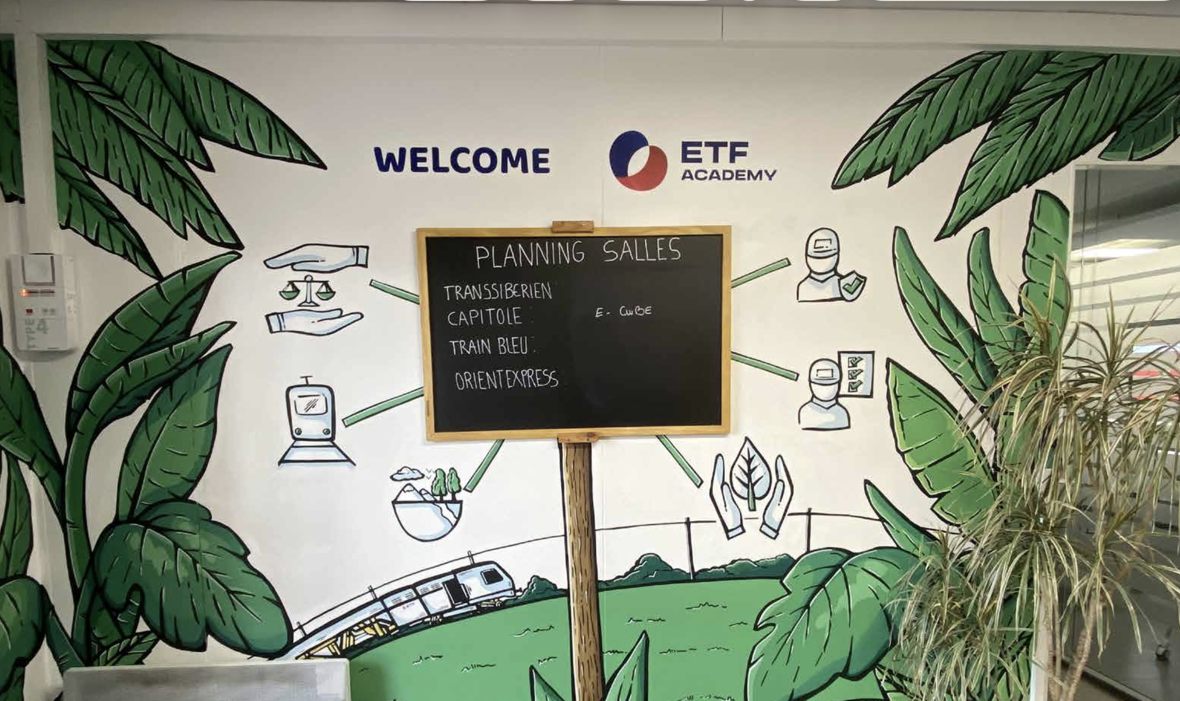 Mur de décoration avec feuilles de bananier, un tableau noir avec planning salles, dessins de personnes avec des casques de protection, une branche de plante verte à droite.