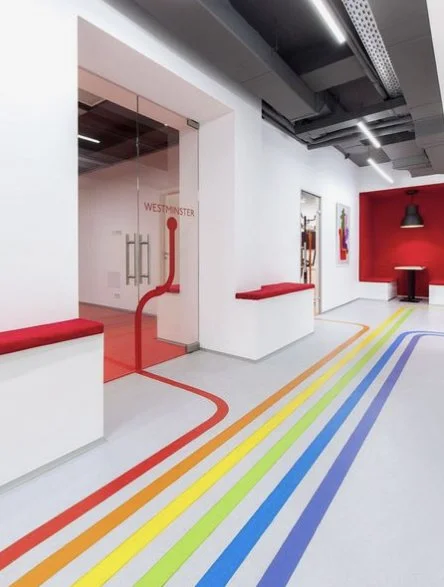 Couloir dans un bureau avec des lignes colorées au sol menant à une porte vitrée, décor moderne avec murs blancs, rouge, et des œuvres d'art.