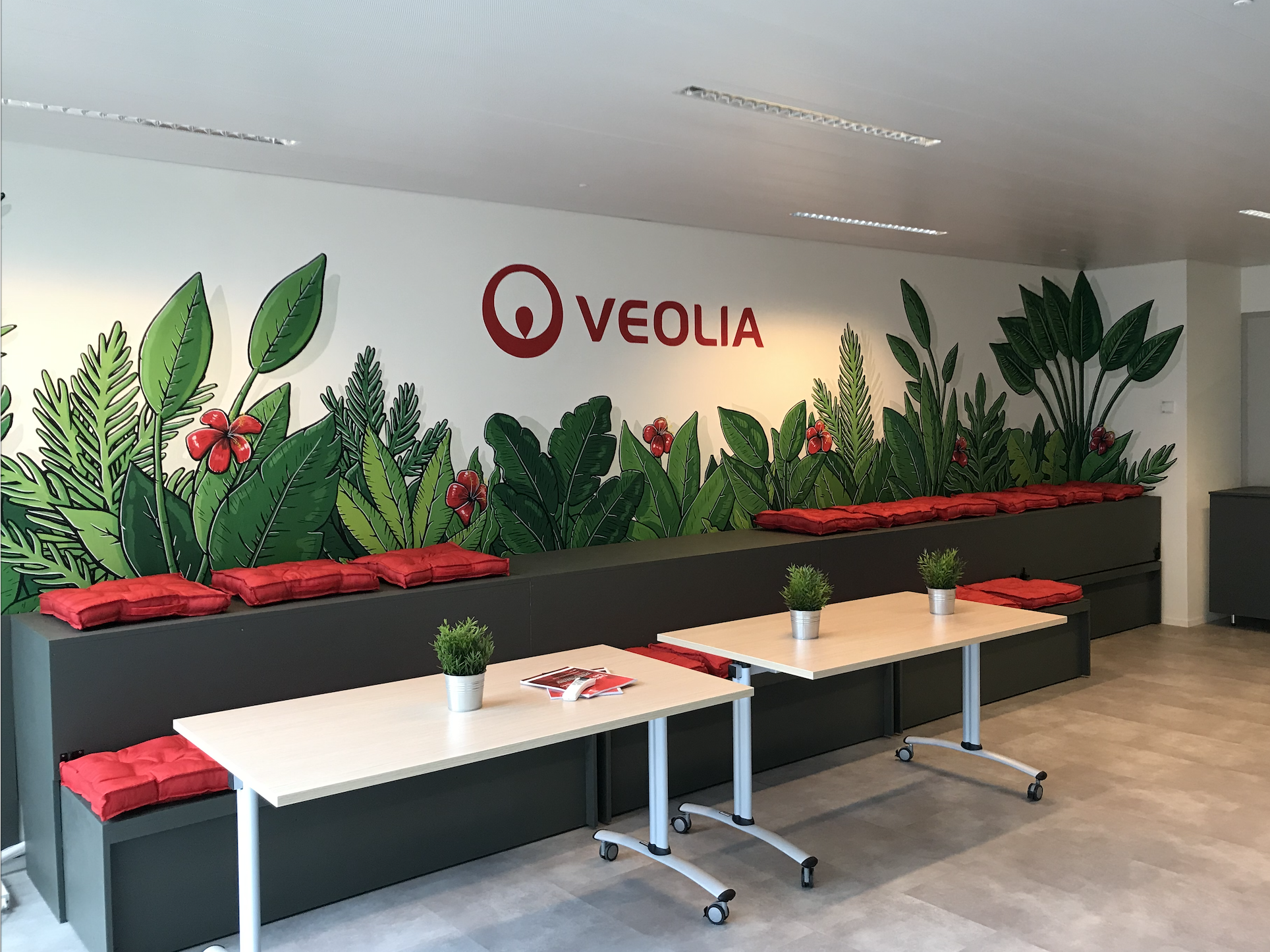 Une salle de réunion avec un mur décoré d'une fresque représentant des feuilles vertes et des fleurs rouges, avec le logo et le nom de Veolia en rouge. Il y a deux tables en bois clair avec des petites plantes en pots, et des coussins rouges disposés le long du mur.