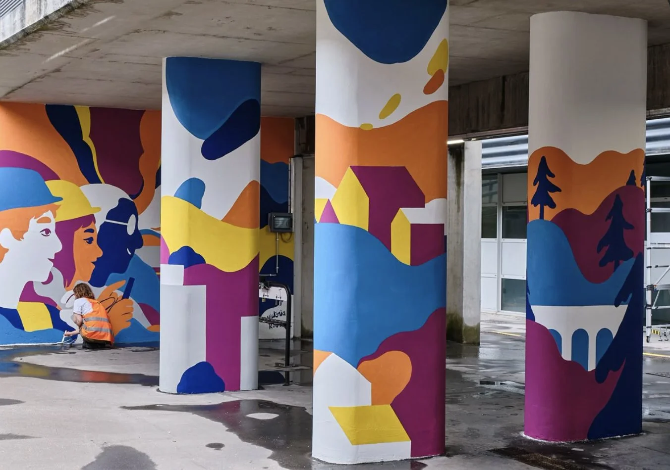 Mural coloré avec des silhouettes de personnes, des maisons, des arbres et des formes abstraites sous un pont