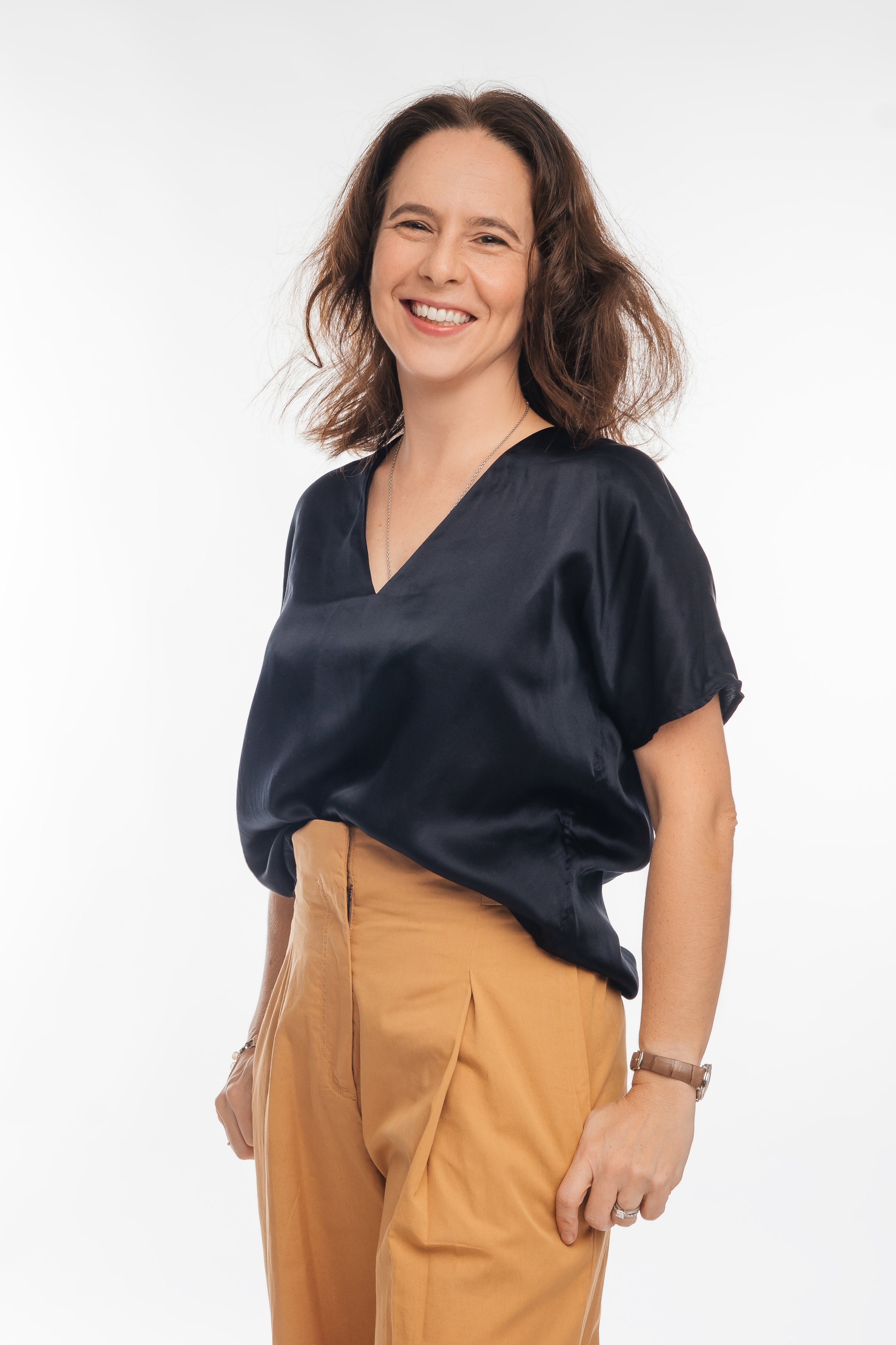 A woman with shoulder-length wavy brown hair, smiling, wearing a black satin blouse and tan high-waisted pants, standing against a plain white background.