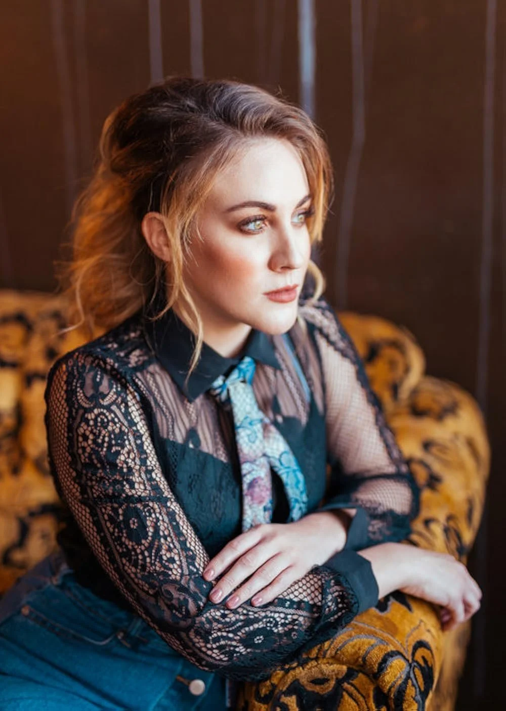 A woman with wavy blonde hair and blue eyes sitting on a vintage sofa with floral upholstery, wearing a black lace top and a colorful scarf.