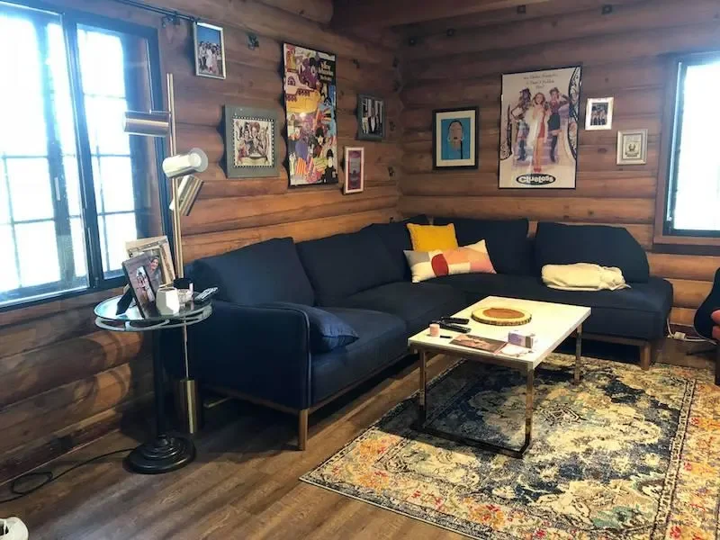 A cozy living room with wooden walls and floors, featuring a large dark blue sectional sofa with colorful pillows. There is a white coffee table with a pizza, remote controls, and candles. Decorative artwork and photos are hung on the wall, and two windows let in natural light.