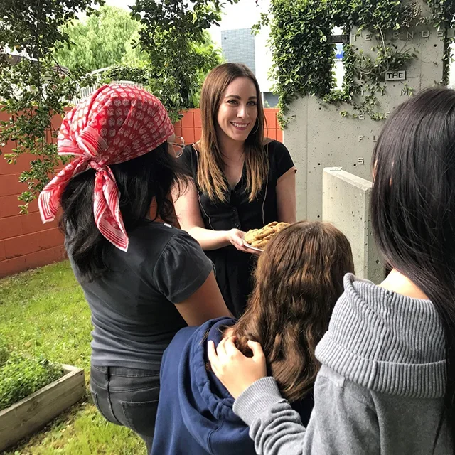 A woman with long brown hair smiling and holding a plate of cookies while talking to a group of four people outside in a backyard garden.