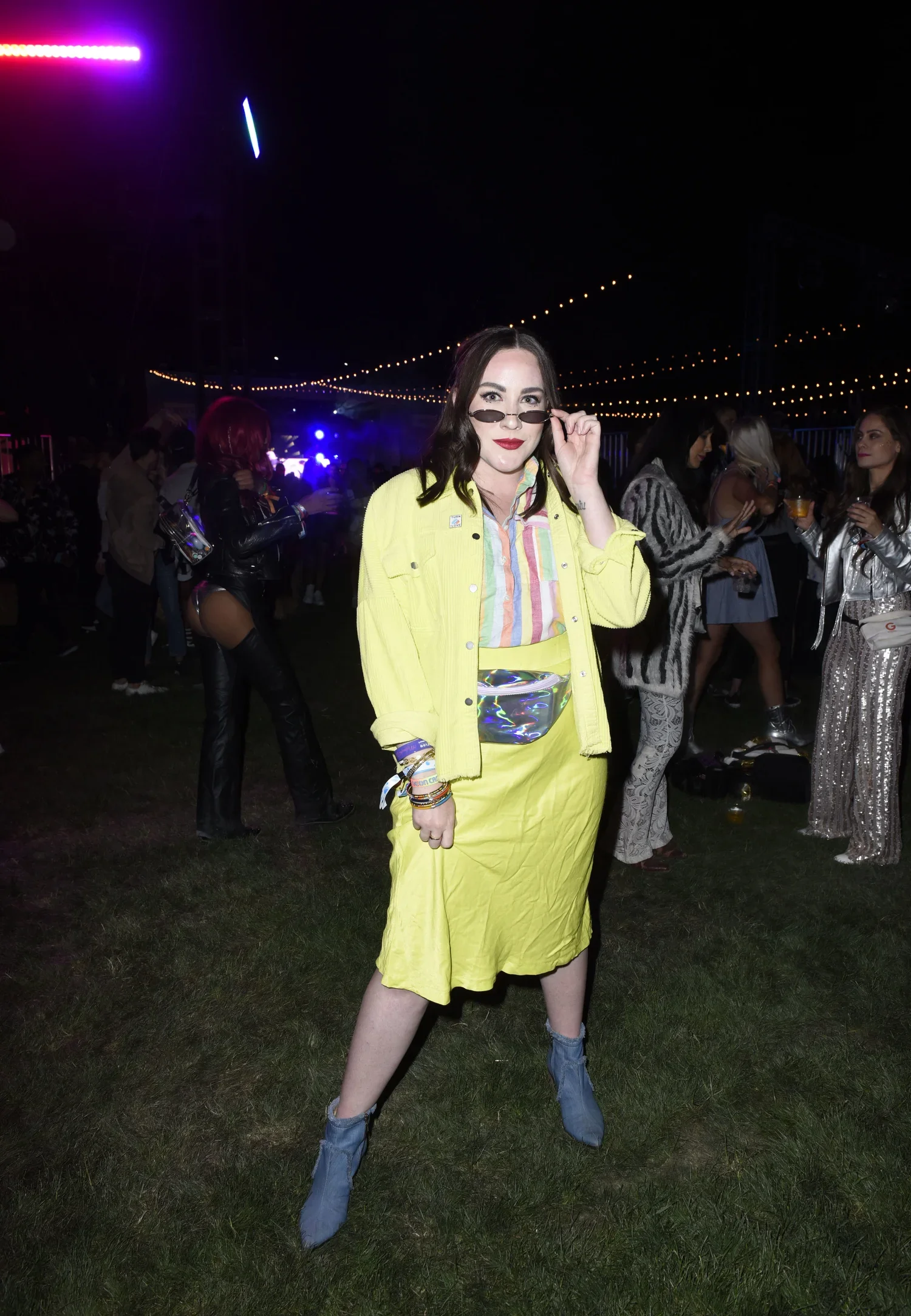 A young woman with dark hair and red lipstick wearing a yellow skirt and jacket poses with sunglasses at a nighttime outdoor event with string lights and people in stylish outfits in the background.