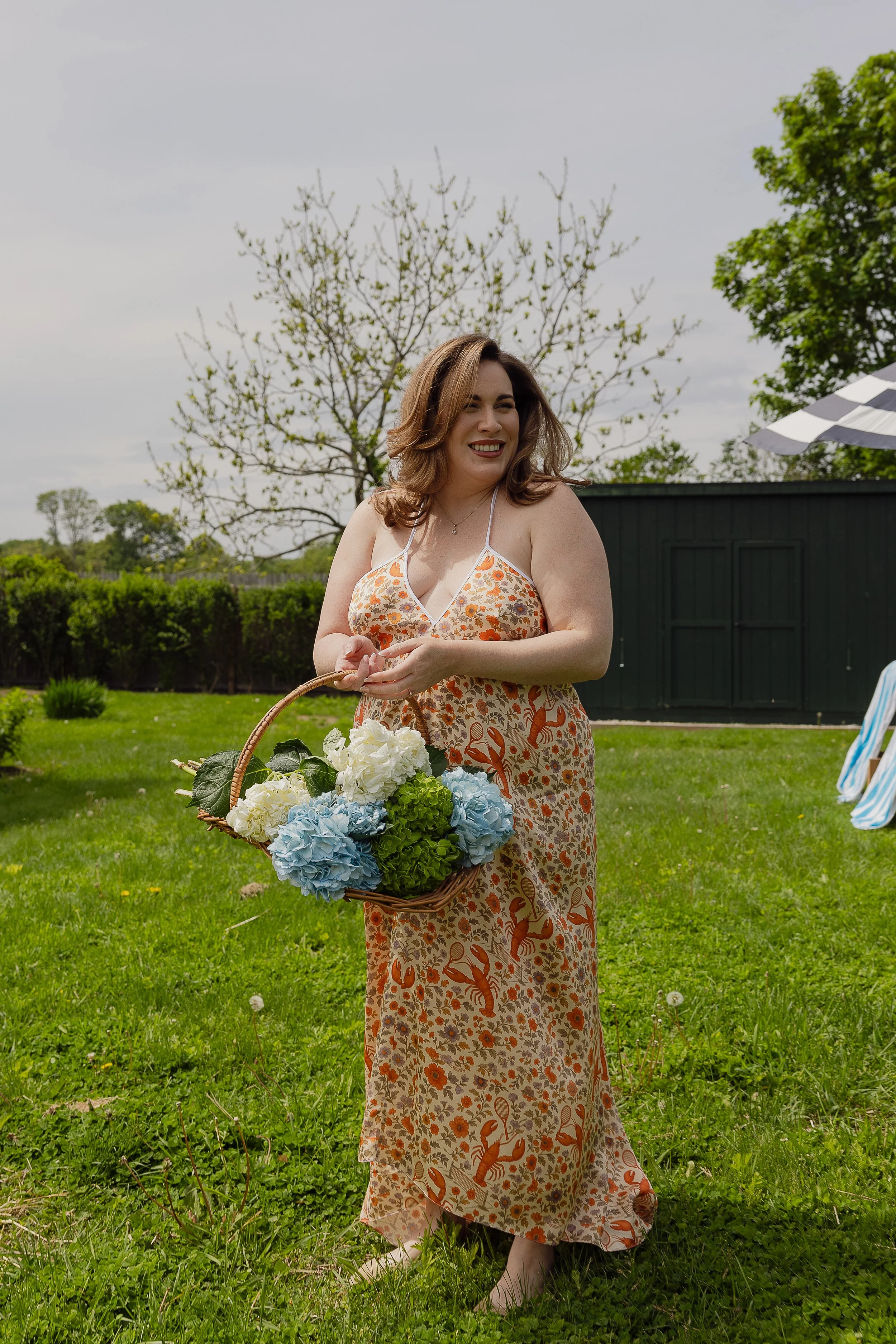 A woman in a floral dress holding a basket of flowers outdoors on a cloudy day, with green grass, trees, and a dark shed in the background.
