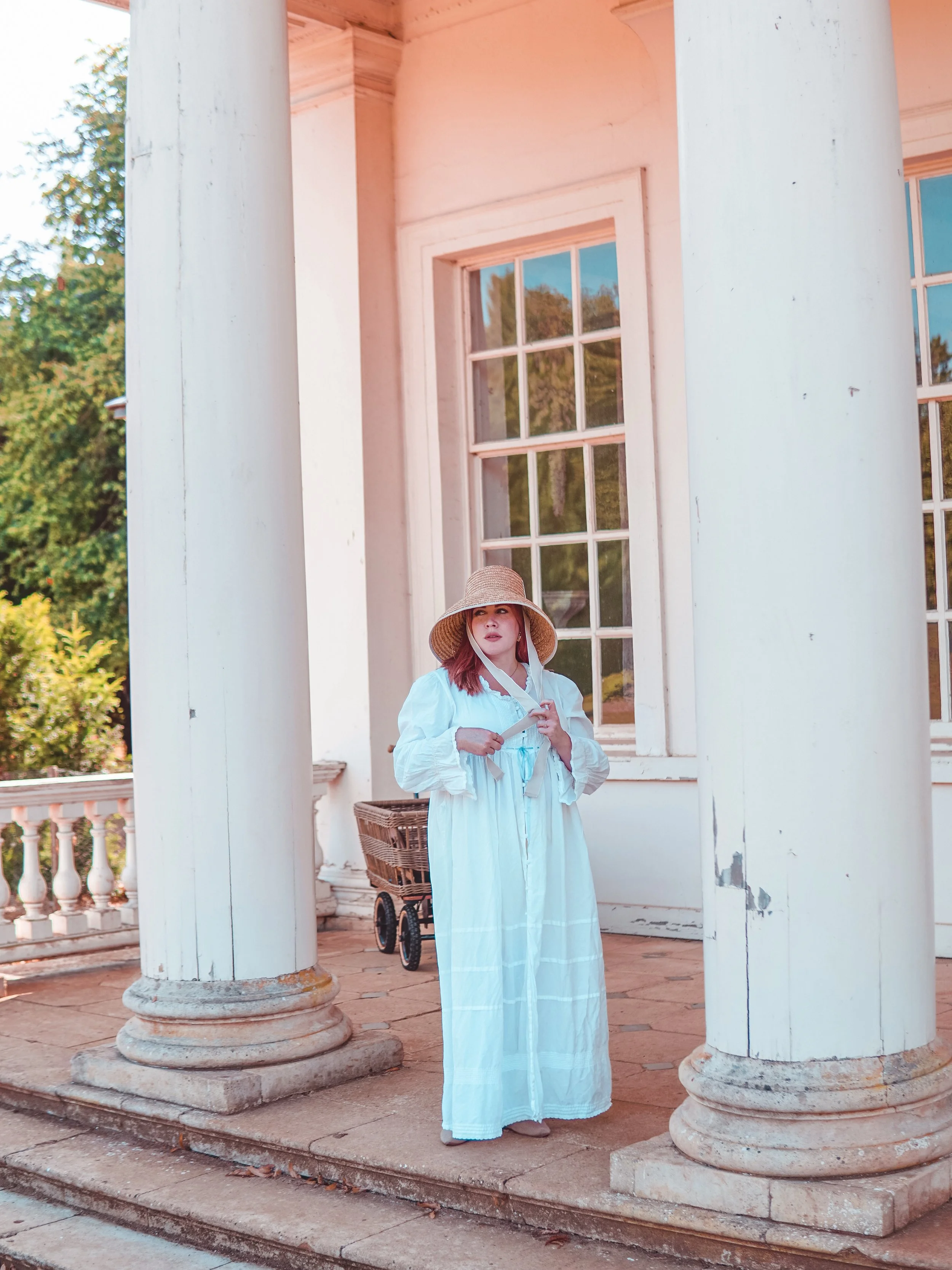 Woman in a white dress and wide-brimmed hat standing on porch of a white wooden house with large windows and columns, carrying a small object in her hands, with a basket or cart behind her.
