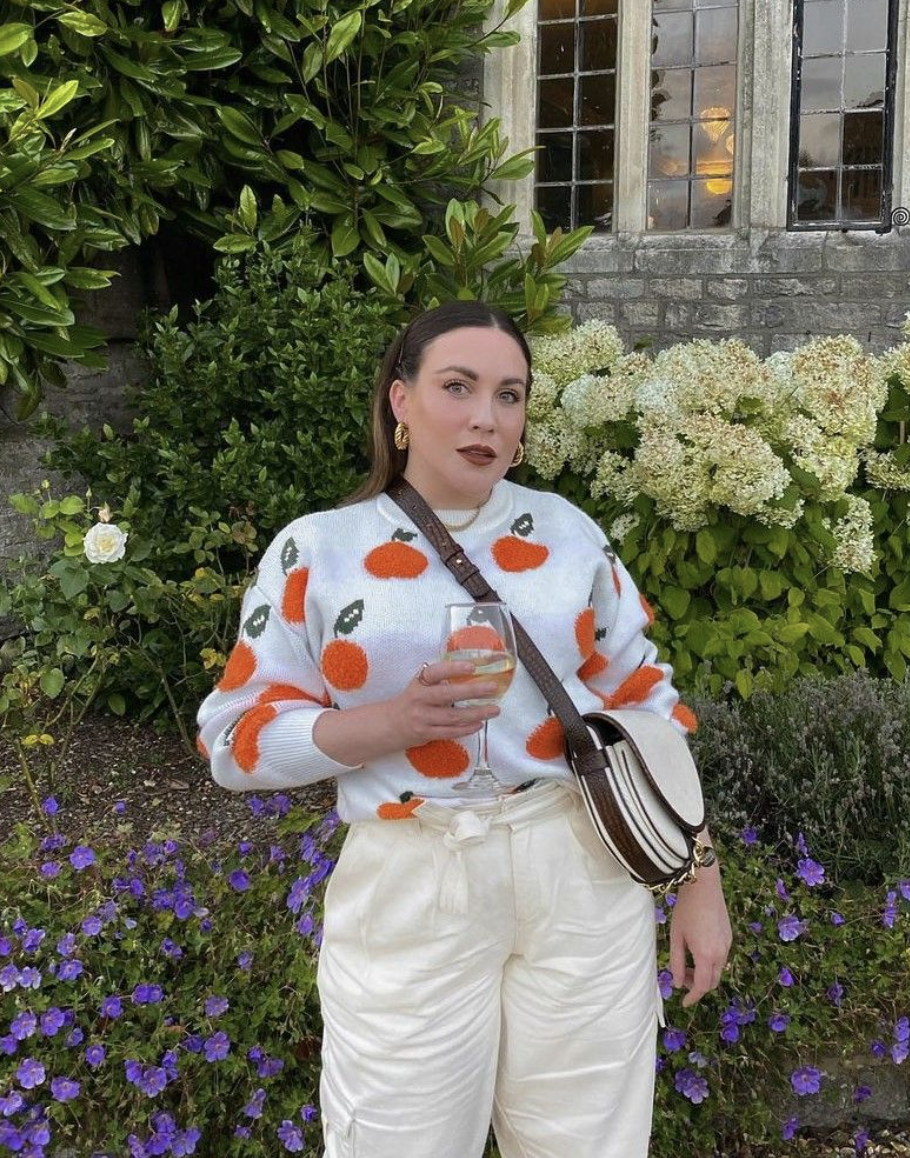 A woman standing in front of a garden with white hydrangeas and purple flowers, holding a glass of white wine, wearing a sweater with orange and green fruit patterns, cream-colored pants, large gold earrings, and a small crossbody bag, next to a rustic brick building with a window.