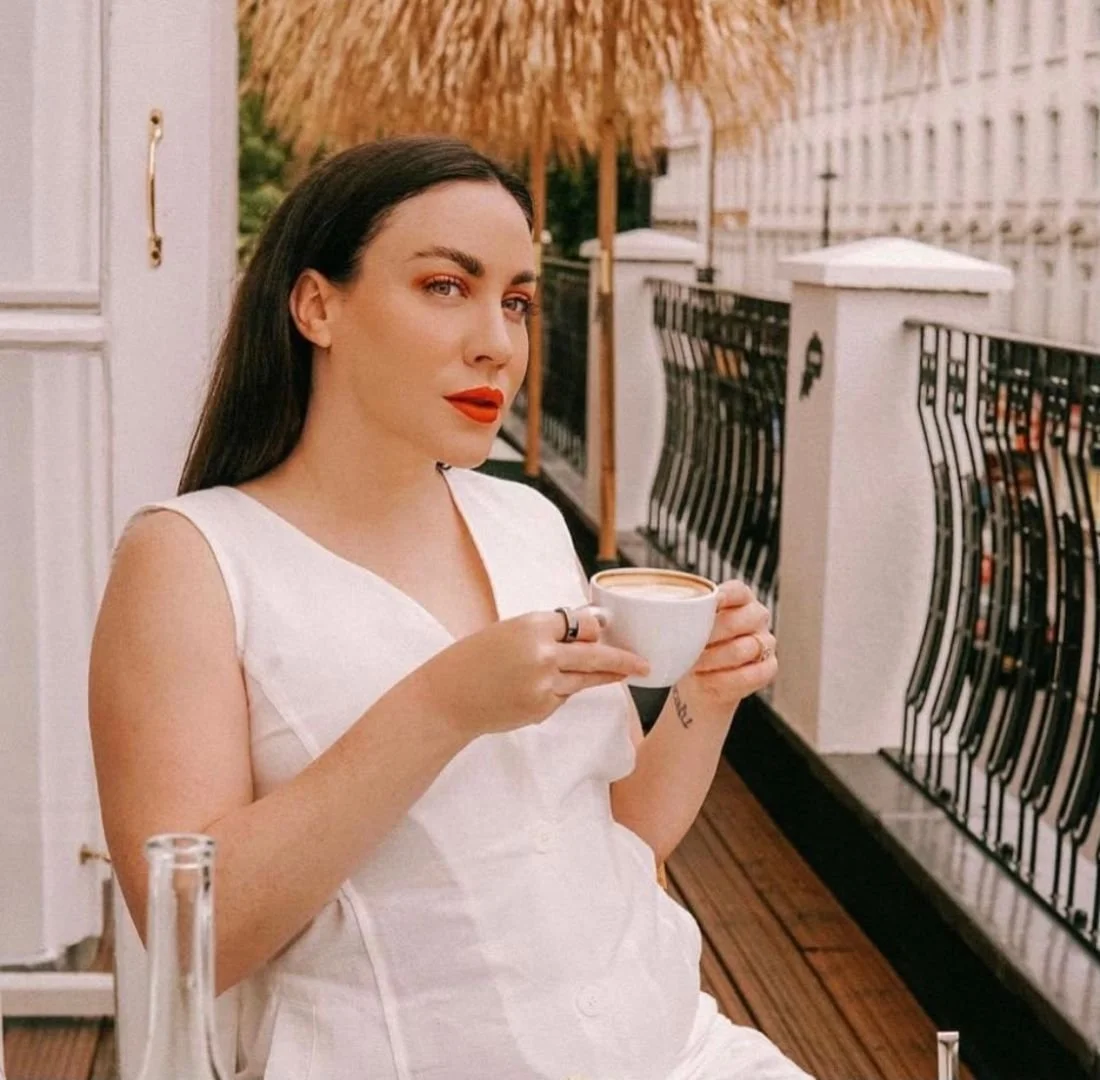 Woman with dark hair and red lipstick sitting outdoors on a balcony, holding a cup of coffee, wearing a white sleeveless top.