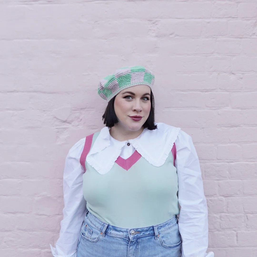 A woman with dark hair and fair skin wearing a pastel green and pink beret, a white blouse, a pastel green vest with pink accents, and high-waisted jeans, standing against a pale pink brick wall.