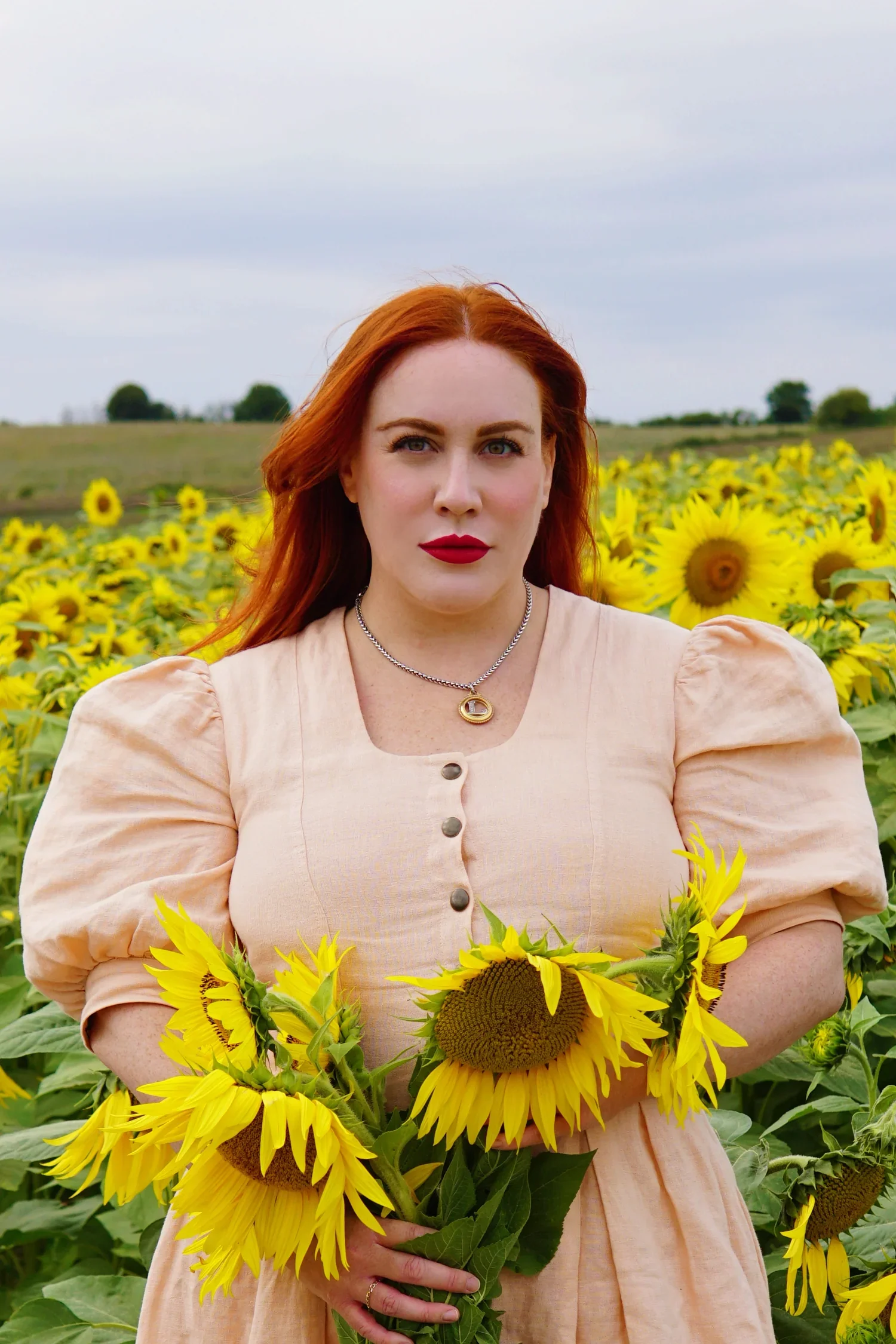 A woman with red hair, wearing a beige dress with puffed sleeves, holding a bunch of sunflowers in a sunflower field.