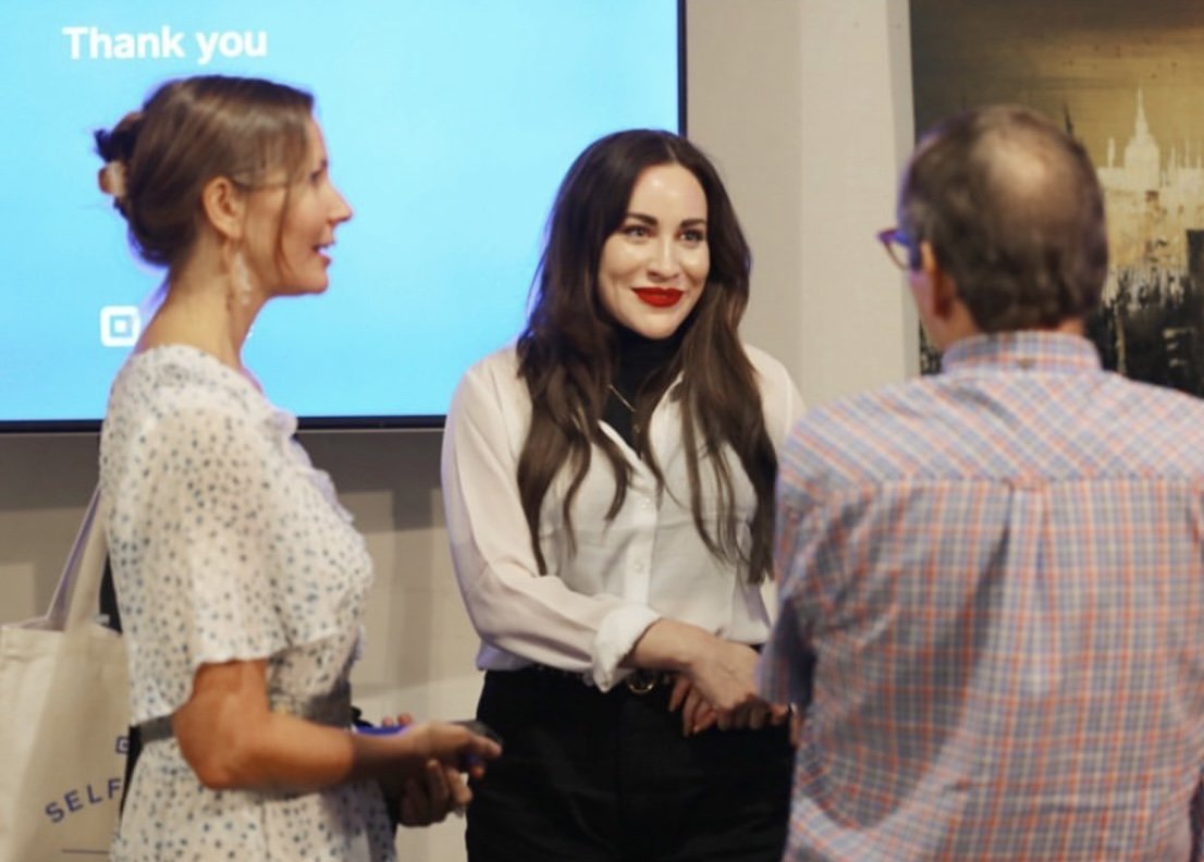 Three people interacting during a professional event, with a woman in the center shaking hands with a man and another woman standing nearby, in front of a blue screen that says 'Thank you'.