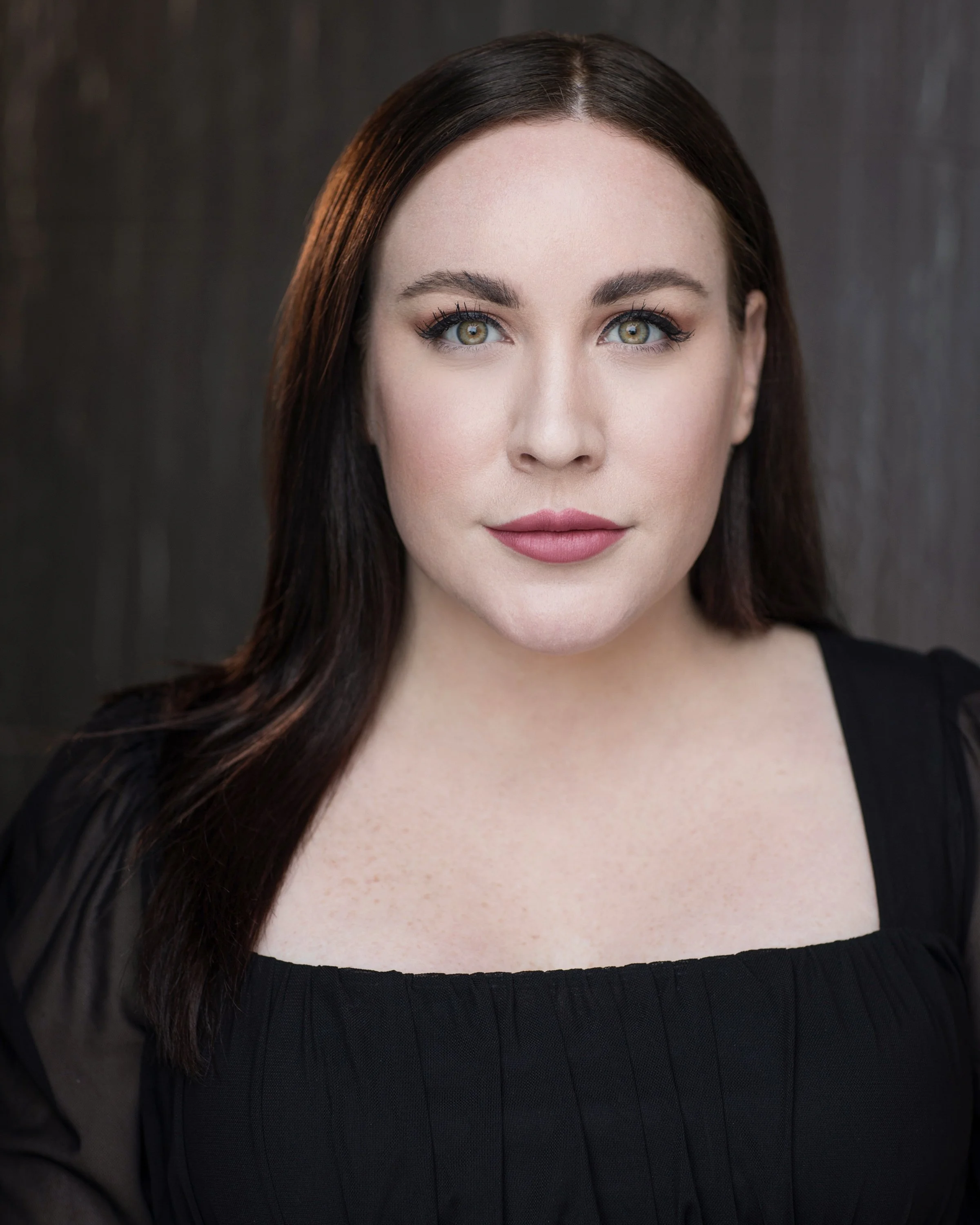 Close-up portrait of a woman with dark brown hair, green eyes, and pink lipstick, wearing a black top with sheer sleeves.