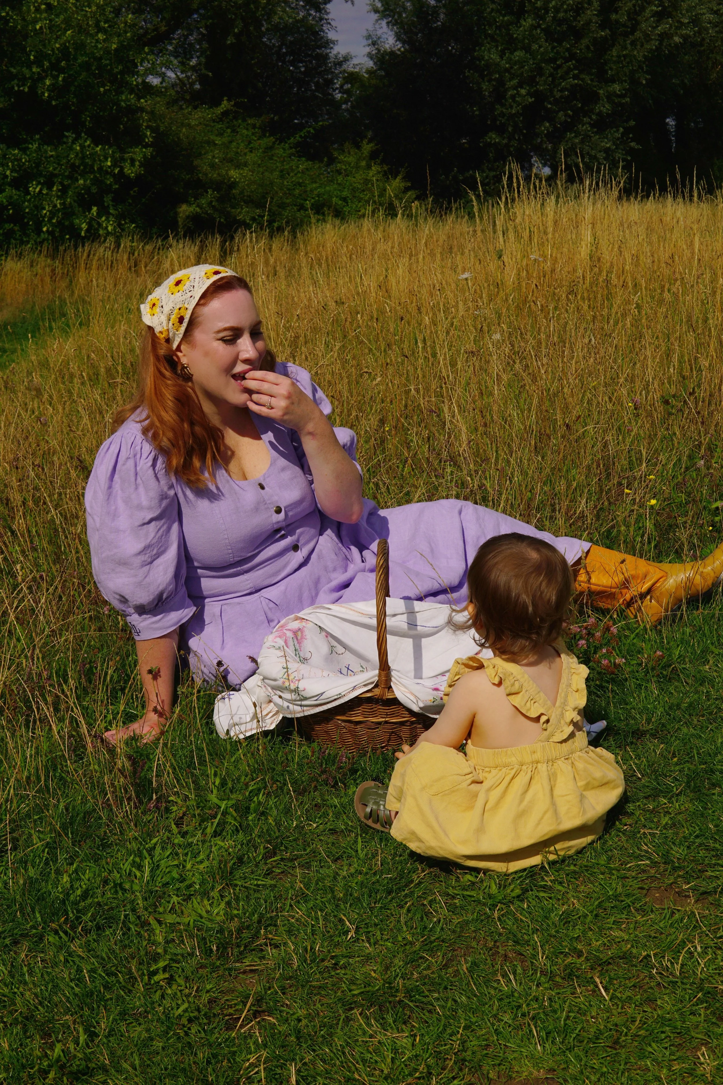 A woman with red hair wearing a purple dress and a sunflower headband sits in a grassy field with a young girl in a yellow dress. They are having a picnic, with a basket and cloth on the grass, surrounded by tall grass and trees in the background.