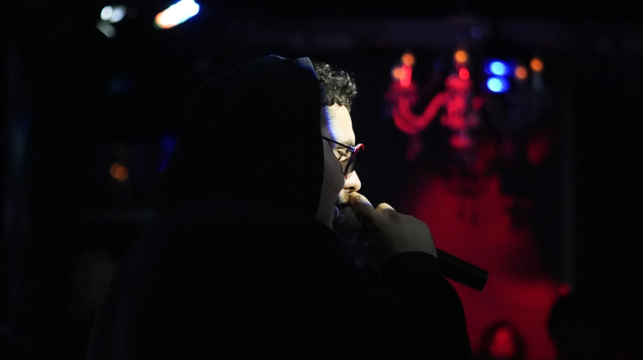 Persona con gafas y gorra negra, sosteniendo un micrófono en un escenario oscuro con luces de colores en el fondo.