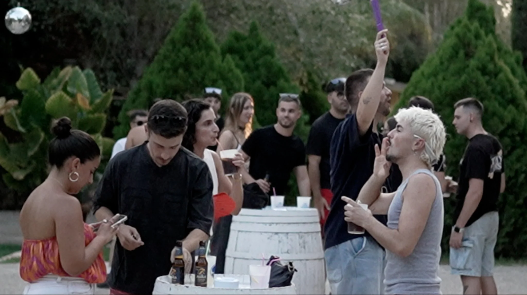 Grupo de personas en una reunión social al aire libre con árboles verdes de fondo, algunos tomando bebidas y otros usando sus teléfonos, mientras una mujer con cabello rubio y cortado en estilo pixie está en conversación cercana con un hombre con gafas y cabello oscuro.