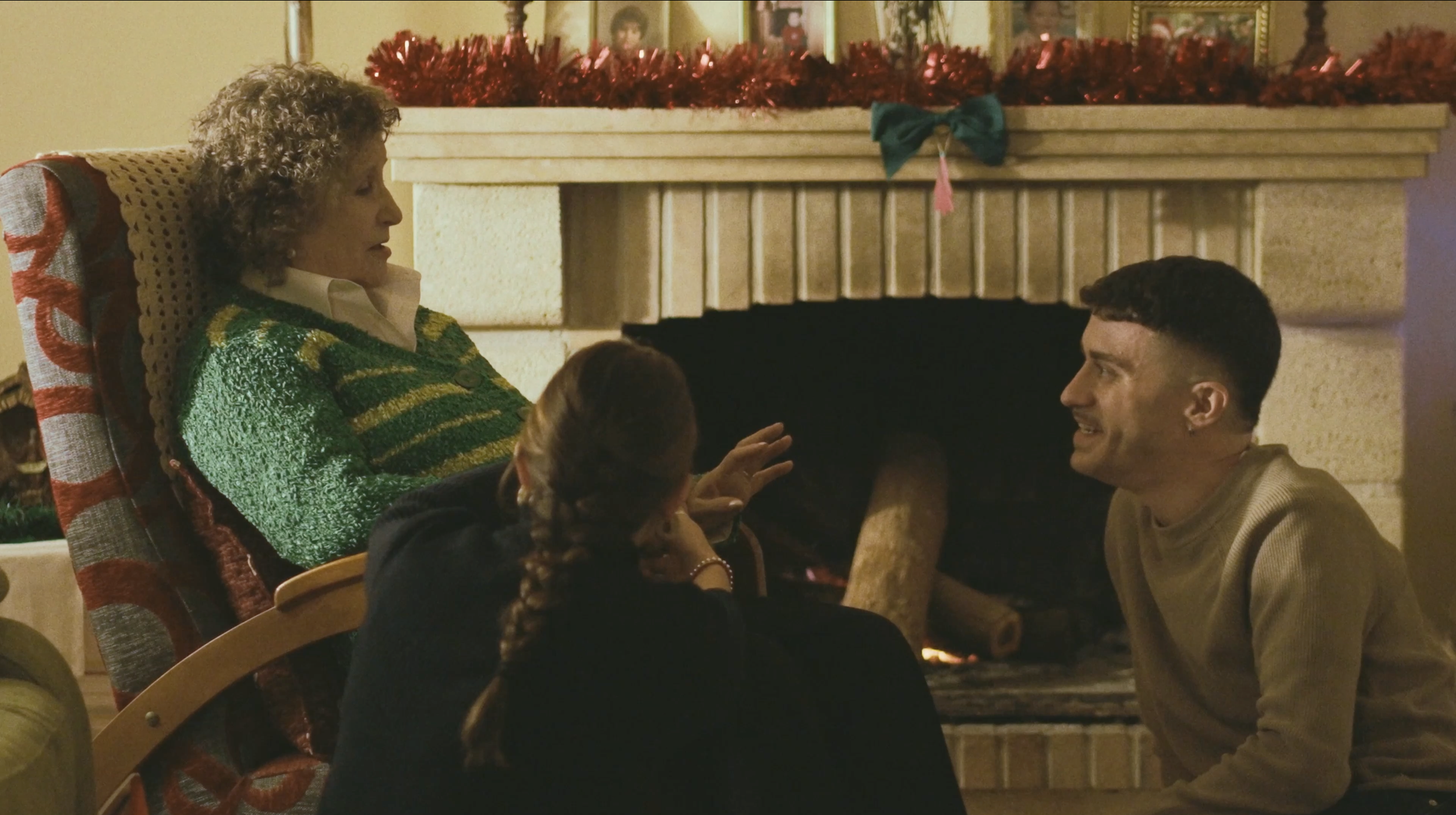 Tres personas en una sala cerca de una chimenea, una mujer mayor en una silla, una joven con trenza y otra mujer joven, conversando con ella durante la Navidad, con adornos navideños en la repisa.