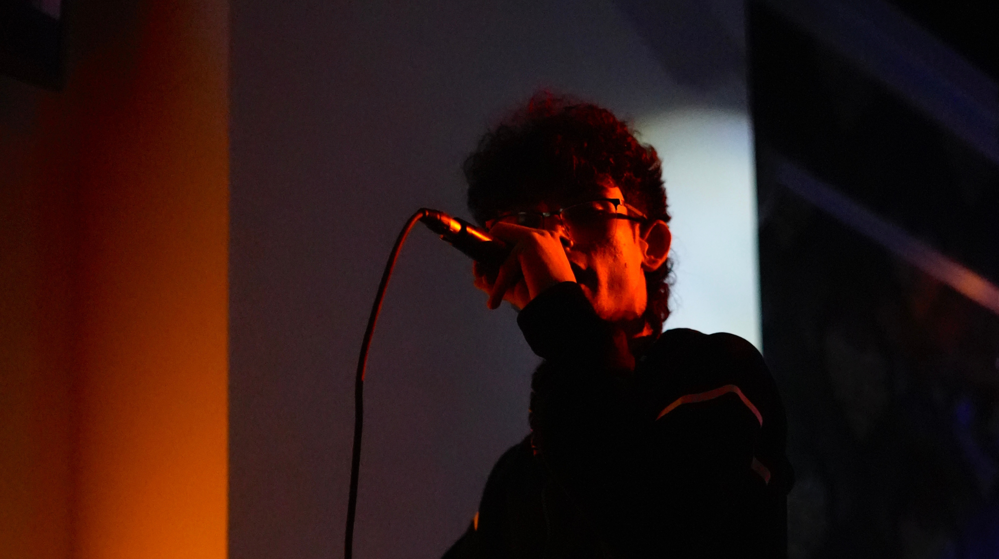 Persona con gafas y cabello rizado sosteniendo un micrófono en un entorno oscuro con iluminación tenue.