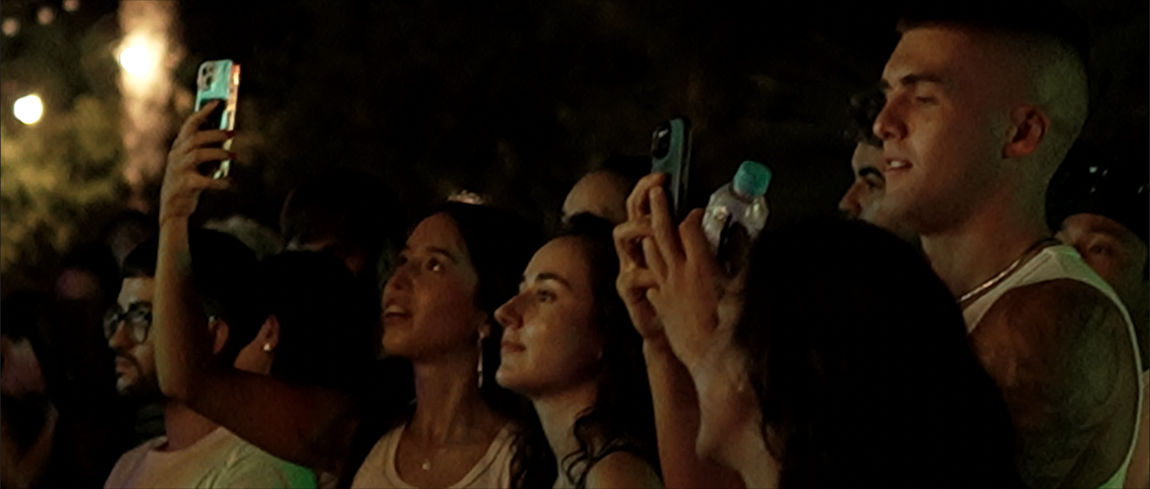 Grupo de personas en un evento nocturno, algunos usando lentes, observando y grabando con sus teléfonos móviles.