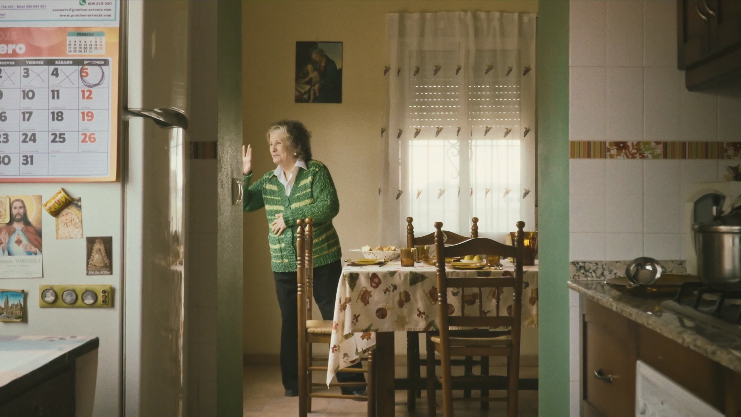 Anciana en la cocina, situada en el comedor, recargada en la puerta, con una mesa decorada con mantelería infantil y comida, luz natural que entra por la ventana con cortinas. En la pared cuadros religiosos.