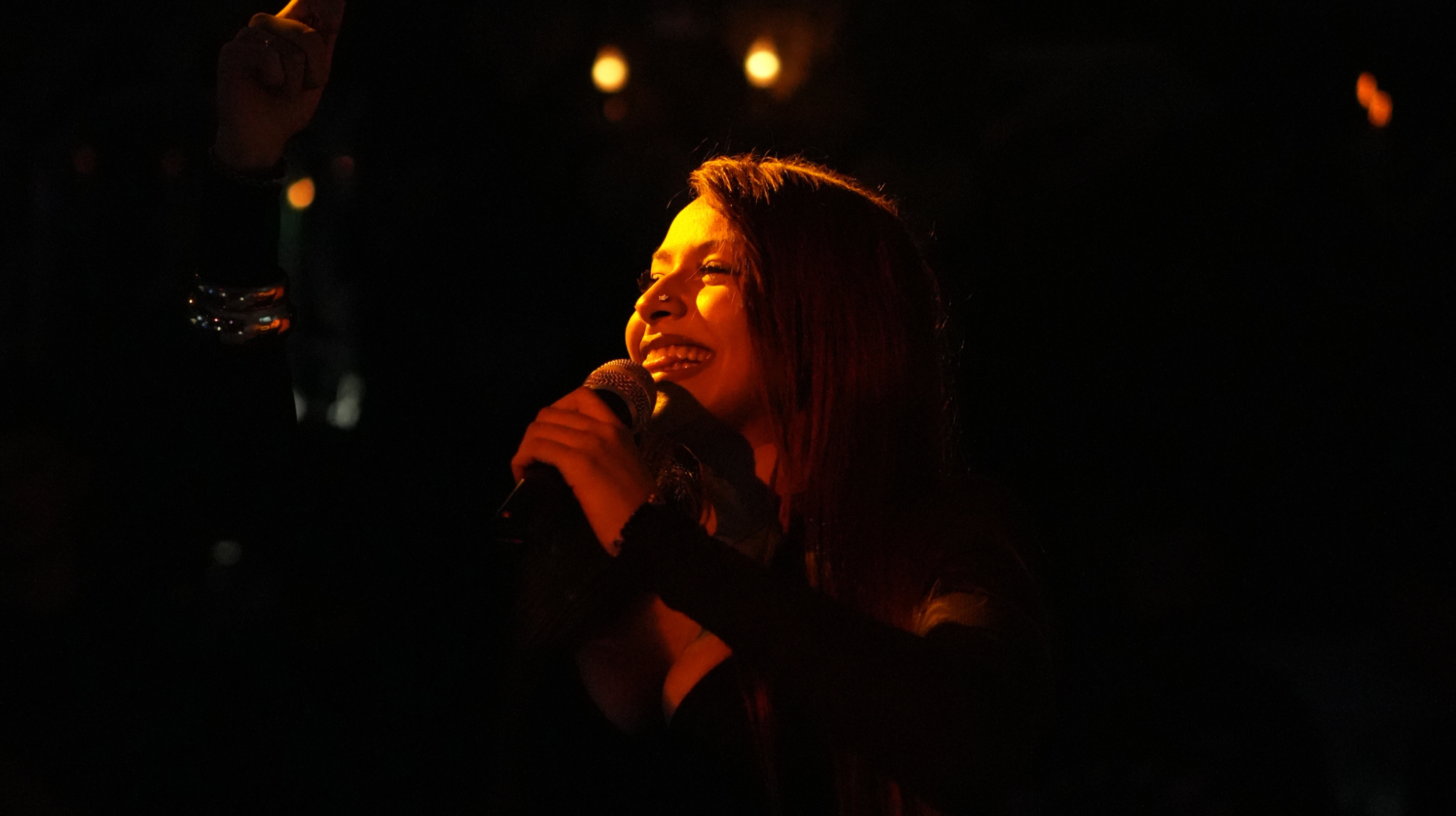 Una mujer feliz con cabello largo entre risas, sosteniendo un micrófono y en un entorno oscuro con luces difusas.