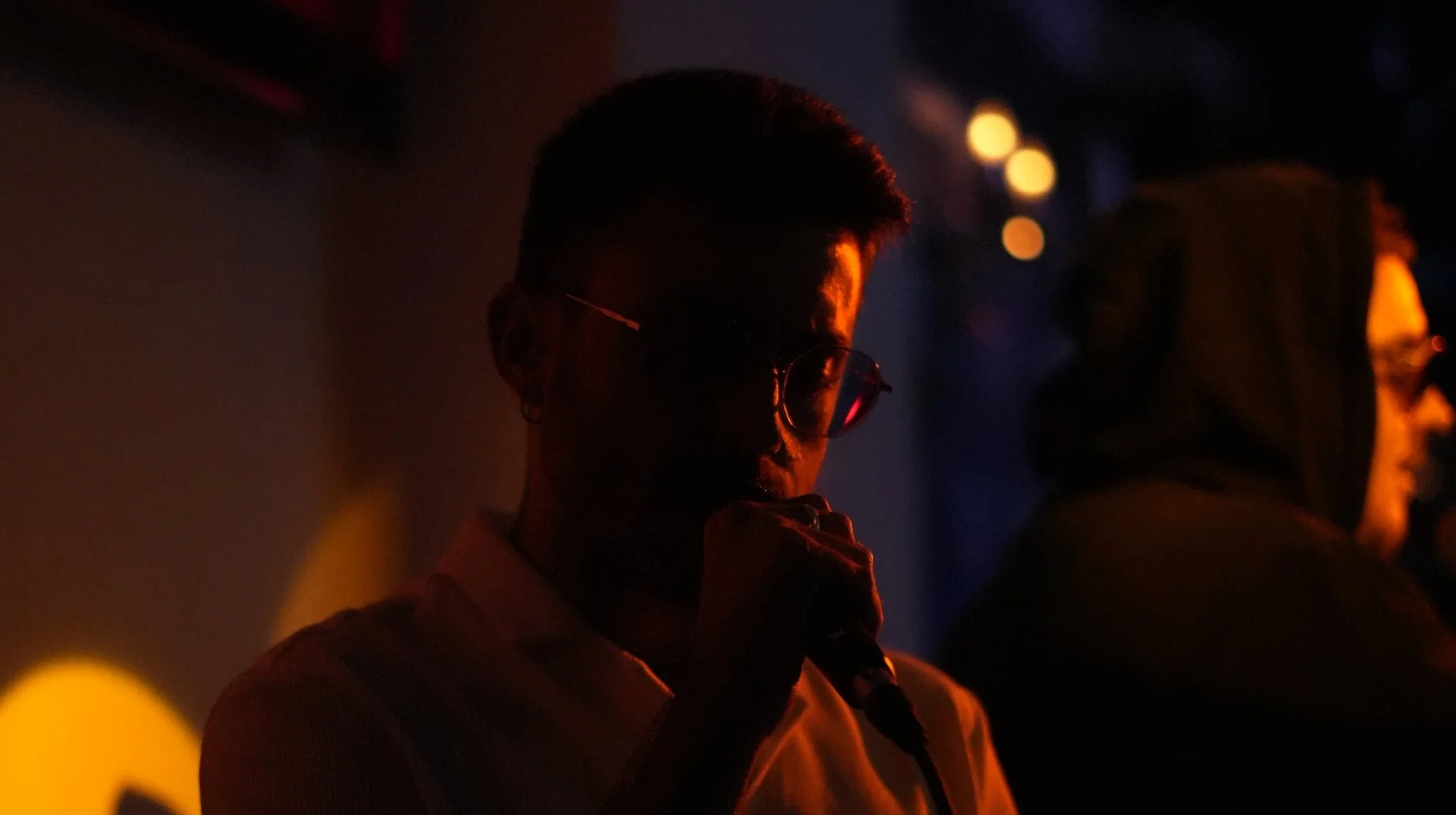 Persona con gafas en un escenario oscuro, sosteniendo un micrófono, iluminada por una luz cálida y tenue, con otra persona en el fondo.