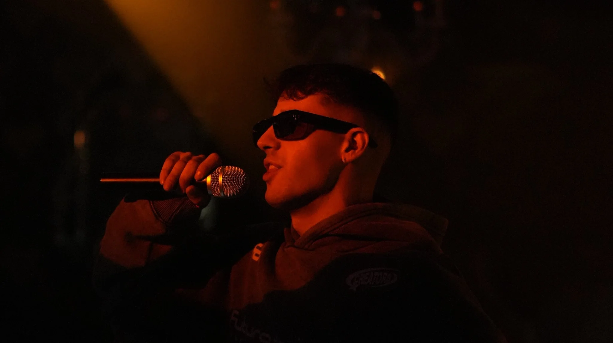 Joven de espaldas en un escenario oscuro con gafas de sol, sosteniendo un micrófono y cantando o hablando para una audiencia