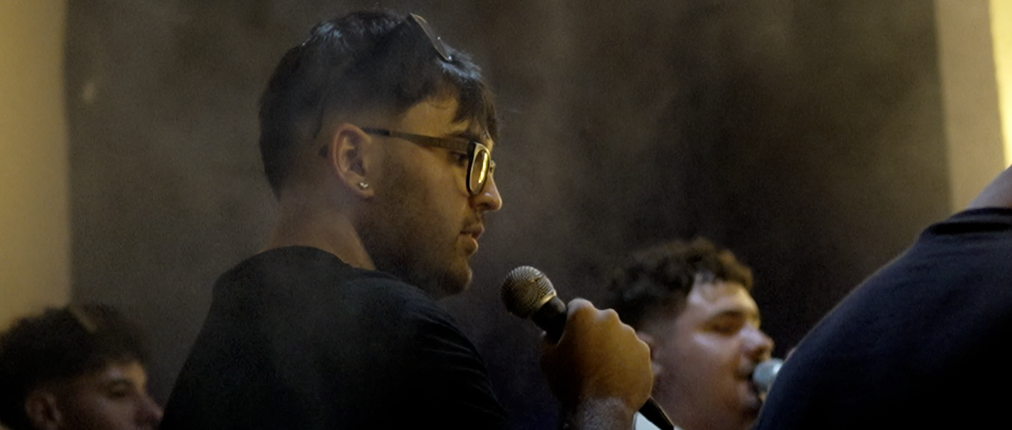 Joven con gafas y barba habla al micrófono en un ambiente oscuro.
