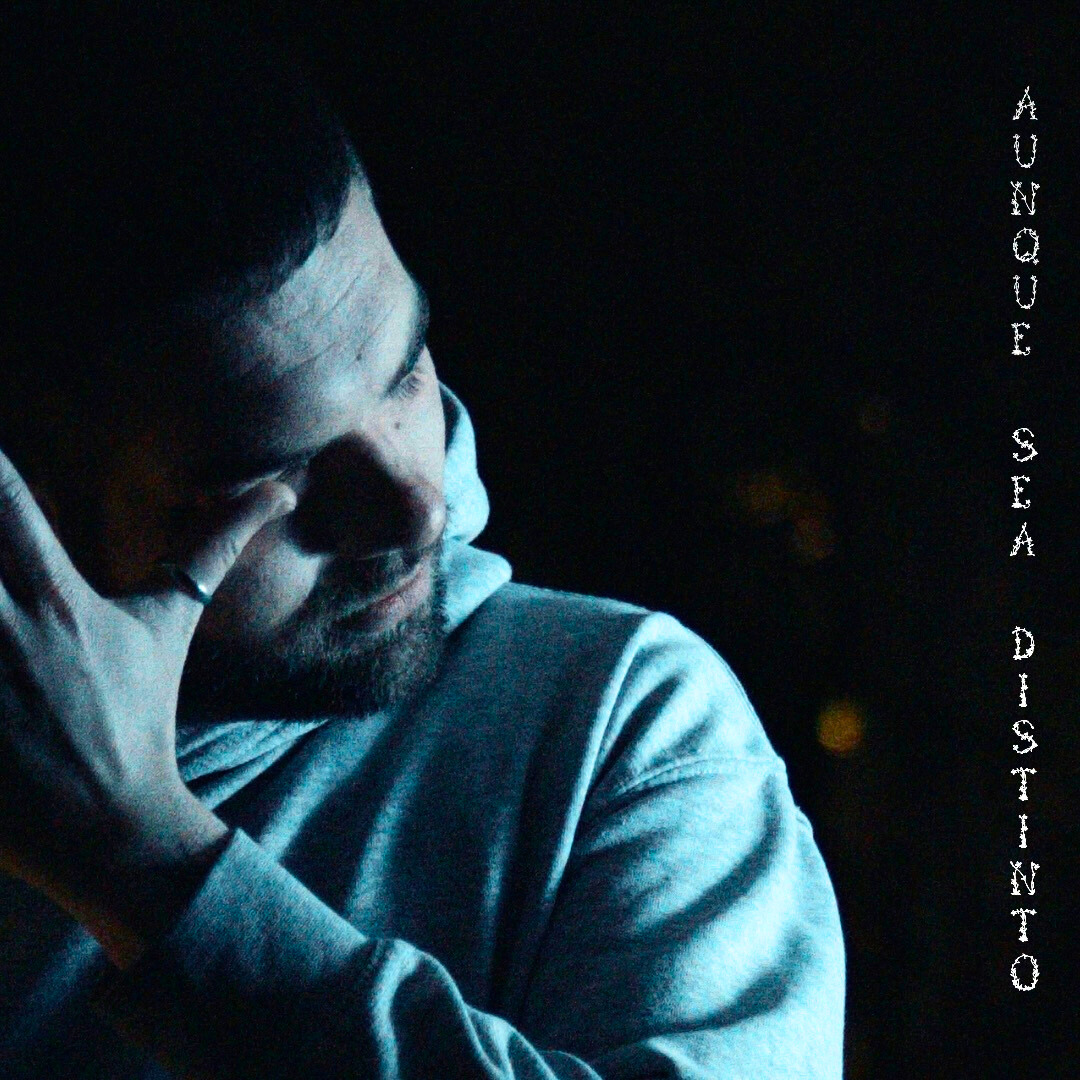 Hombre en una expresión pensativa en un entorno oscuro, con una mano en la cara, vistiendo ropa gris y una camiseta blanca.