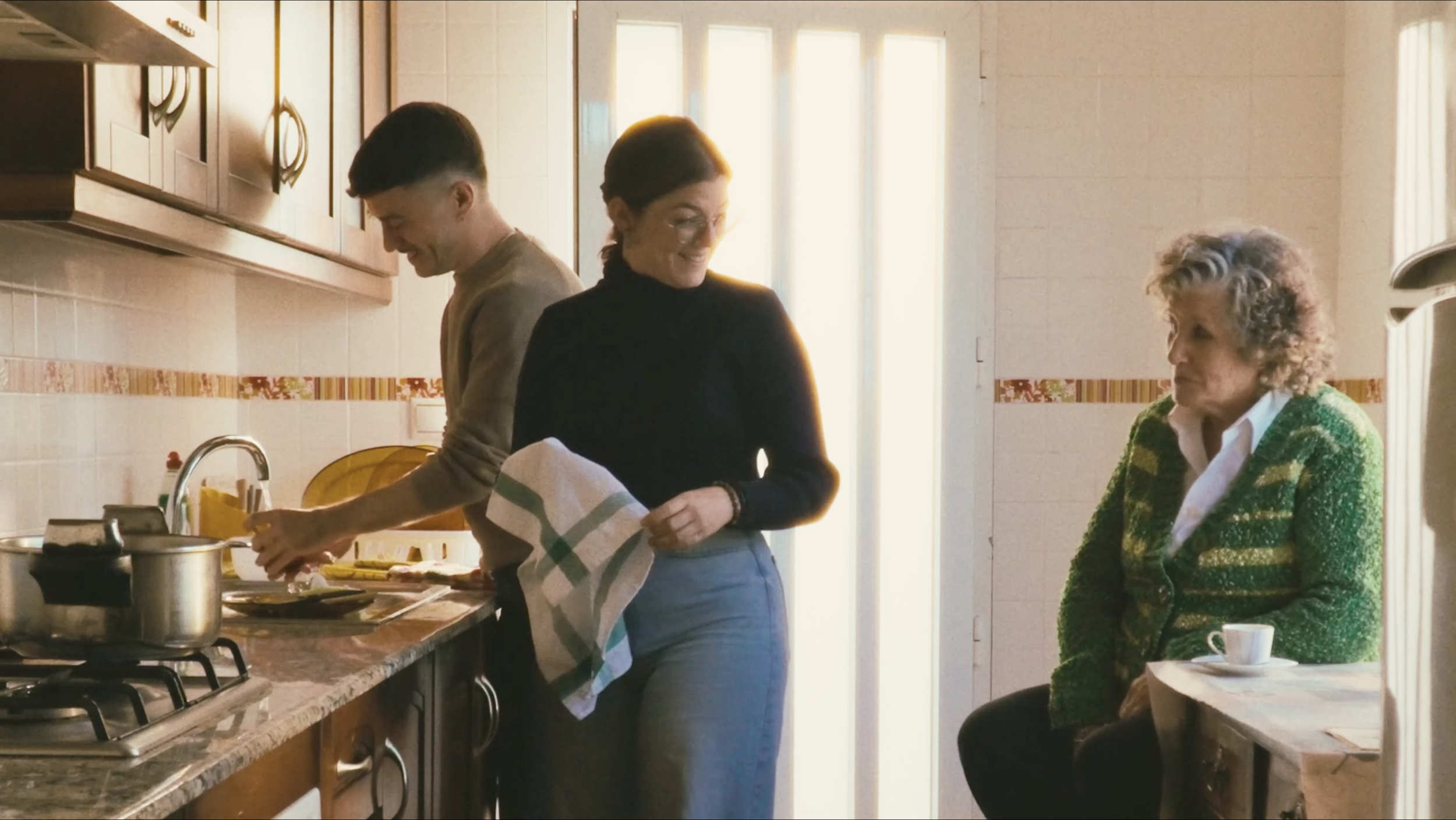 Tres personas en una cocina: un joven y una mujer en la estufa cocinando, una mujer mayor sentada en un banco con una taza de café viendo, todos parecen disfrutar del momento