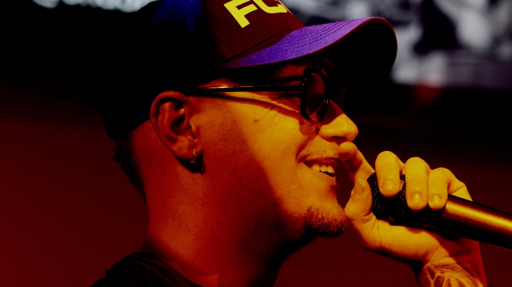 Joven cantando en un micrófono, lleva gafas, gorra roja con azul, e oreja con pendiente.