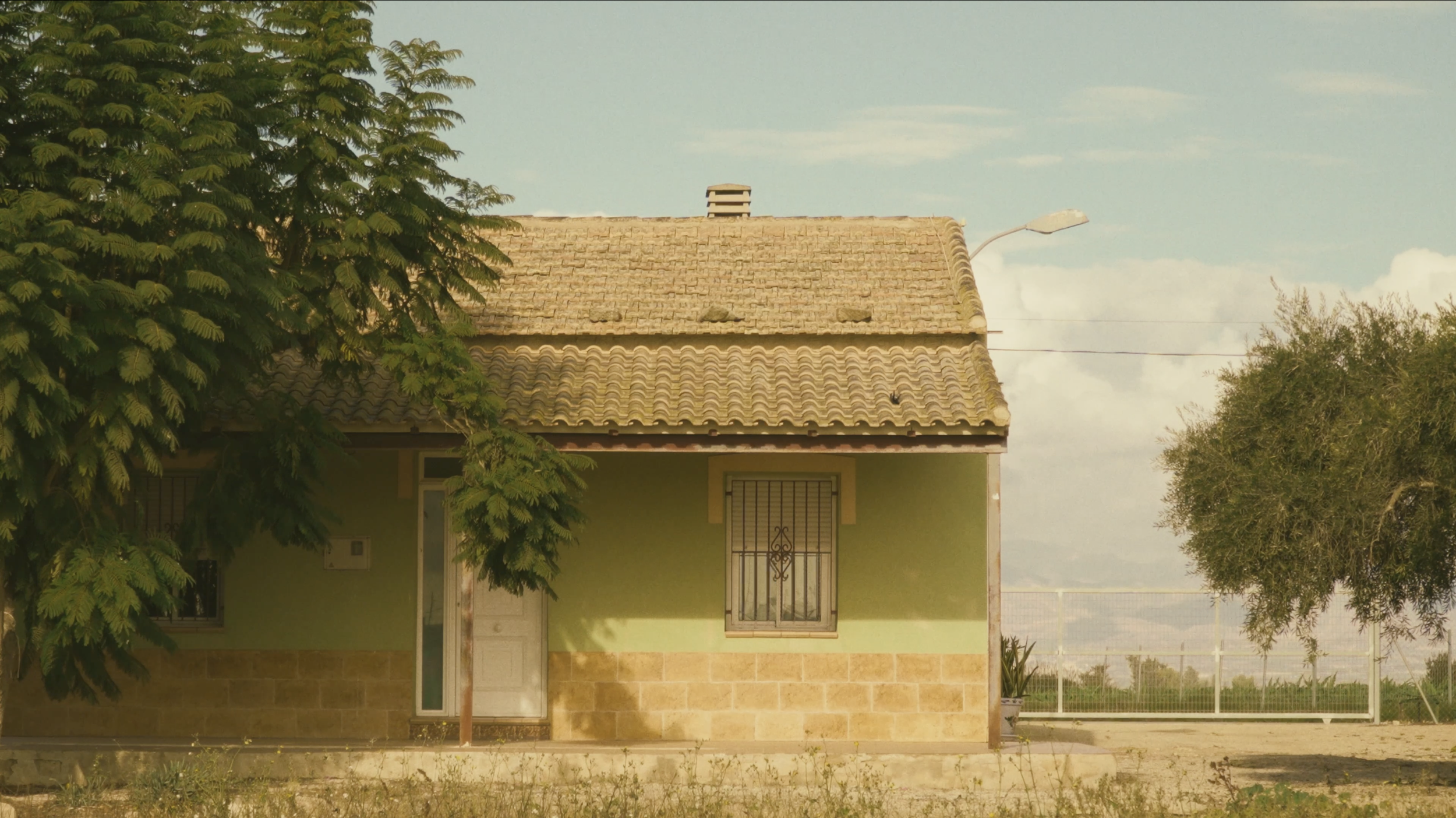 Casa pequeña con pared de ladrillo en la parte inferior, puerta blanca y ventanas con rejas negras, en el frente hay árboles verdes y césped, con cielo parcialmente nublado.