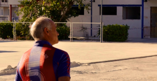 Persona mayor de espaldas en un campo de fútbol, con uniforme de deporte, en un entorno urbano durante el día.